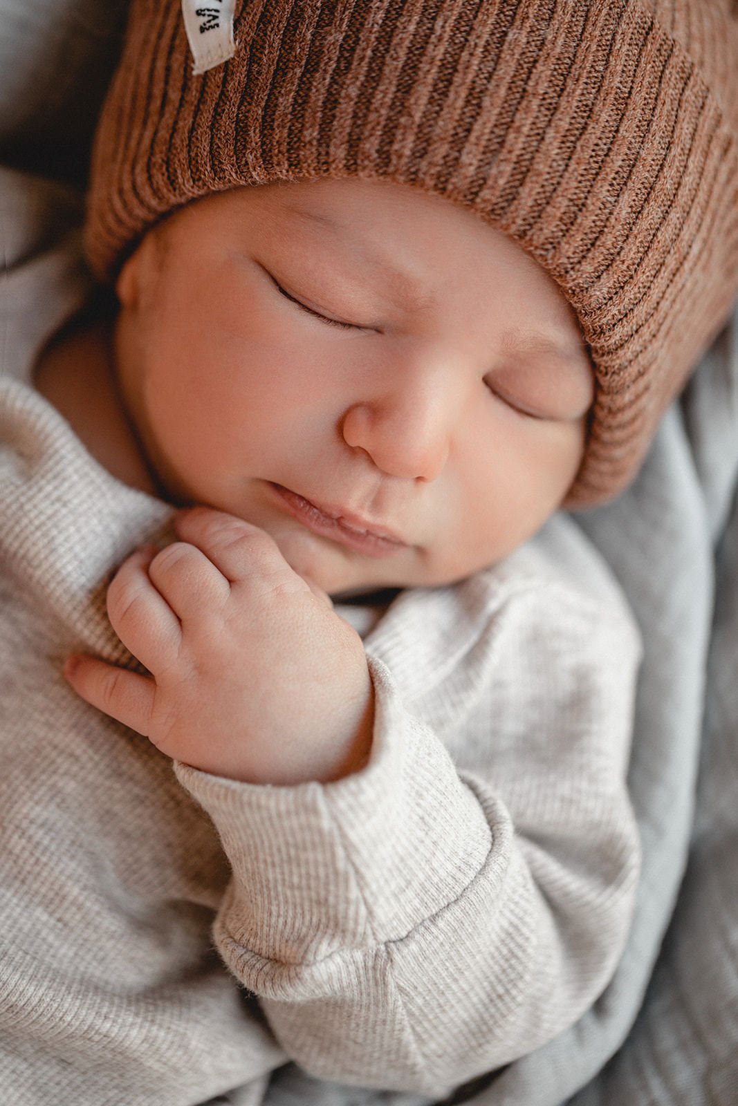 Nahaufnahme eines schlafenden Babys mit einem braunen, gestreiften Mützchen, das eine cremfarbene Sweatjacke trägt.
