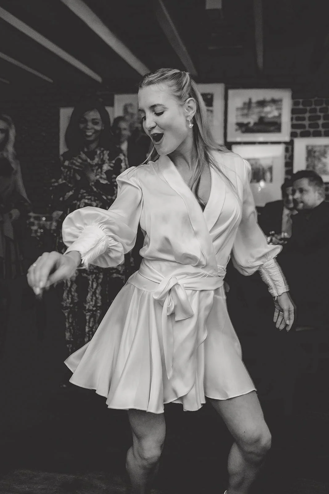 Eine Frau tanzt in einem Kleid in einem Raum mit mehreren aufmerksamen Zuschauern, im Hintergrund hängen Bilder. Sie sieht glücklich aus.