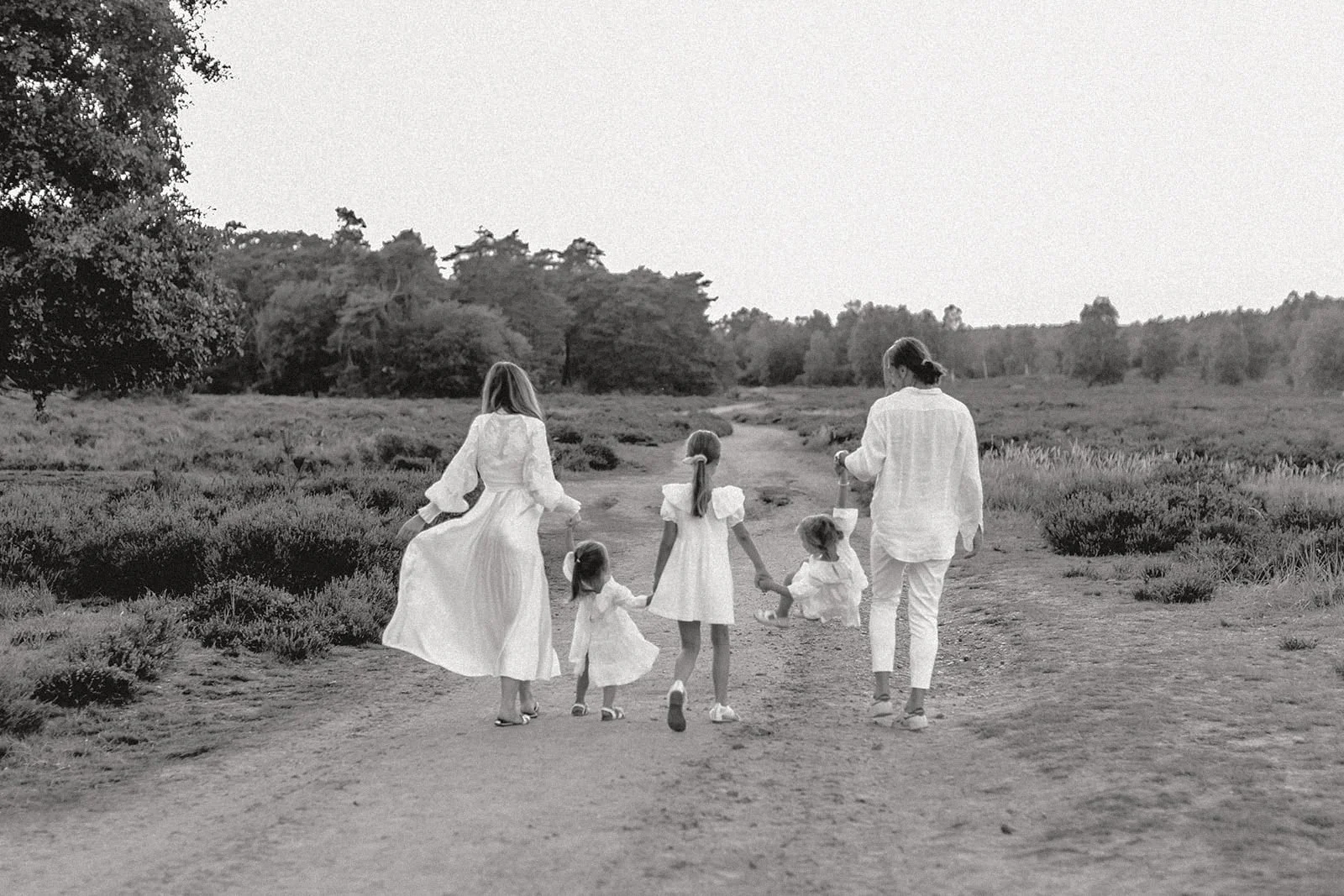Familie mit fünf Kindern geht Hand in Hand auf einem Feldweg im Freien, im Hintergrund Bäume, alles in schwarz-weiß aufgenommen.