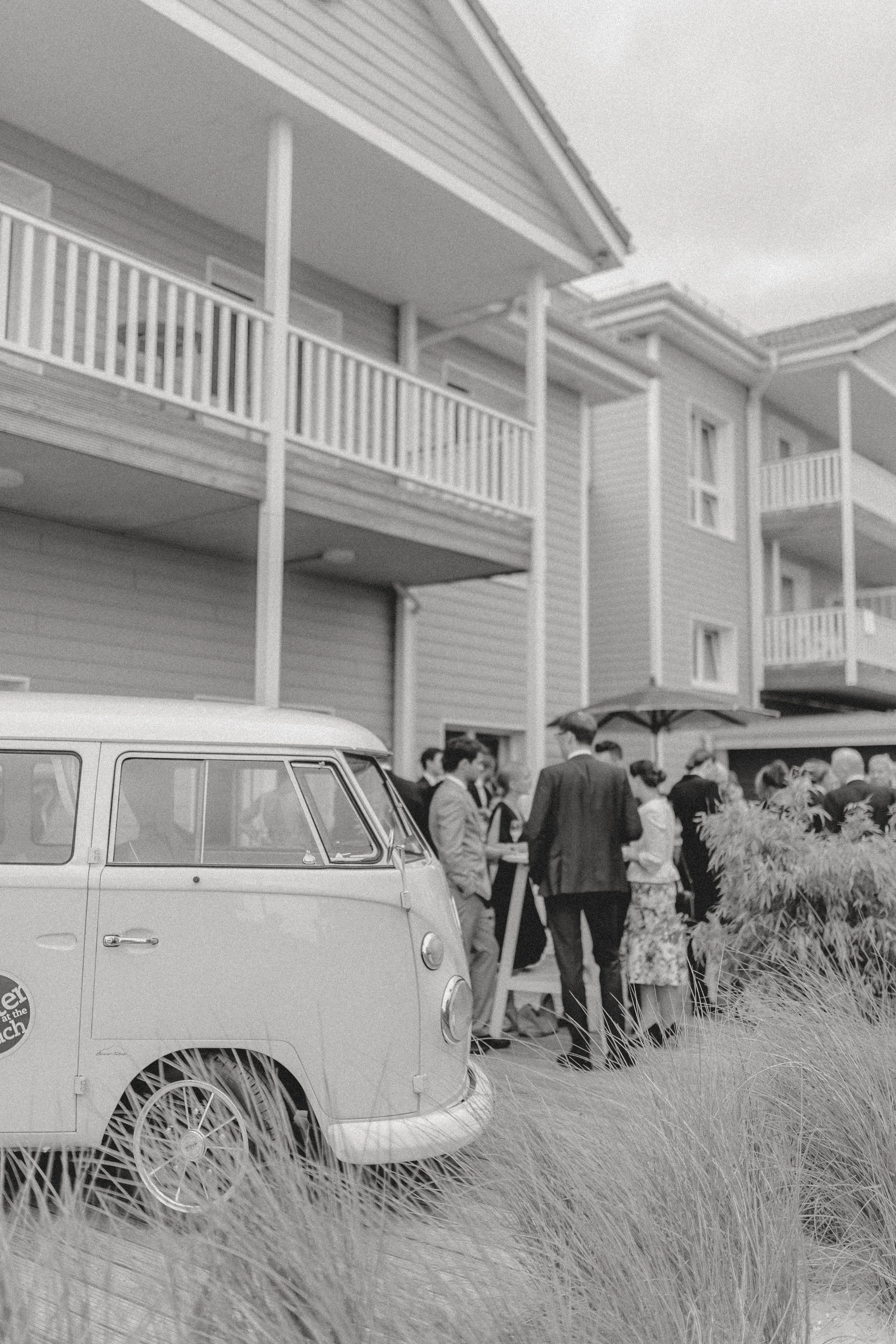 Ein schwarzer und weißer vintage VW-Bus vor einem Mehrfamilienhaus mit mehreren Balkonen; davor stehen Menschen bei einer Veranstaltung im Freien, einige im Gespräch, unter einem Sonnenschirm.