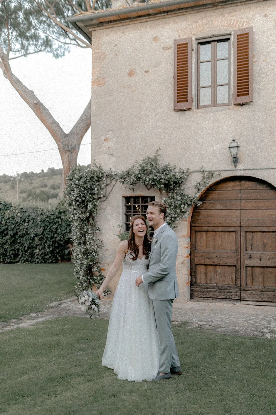 Ein glückliches Brautpaar lacht und tanzt auf dem Rasen vor einem alten Steinhaus mit Holztür, Fenster mit Holzläden und einer Weinrebe an der Wand, im Freien bei Sonnenuntergang.