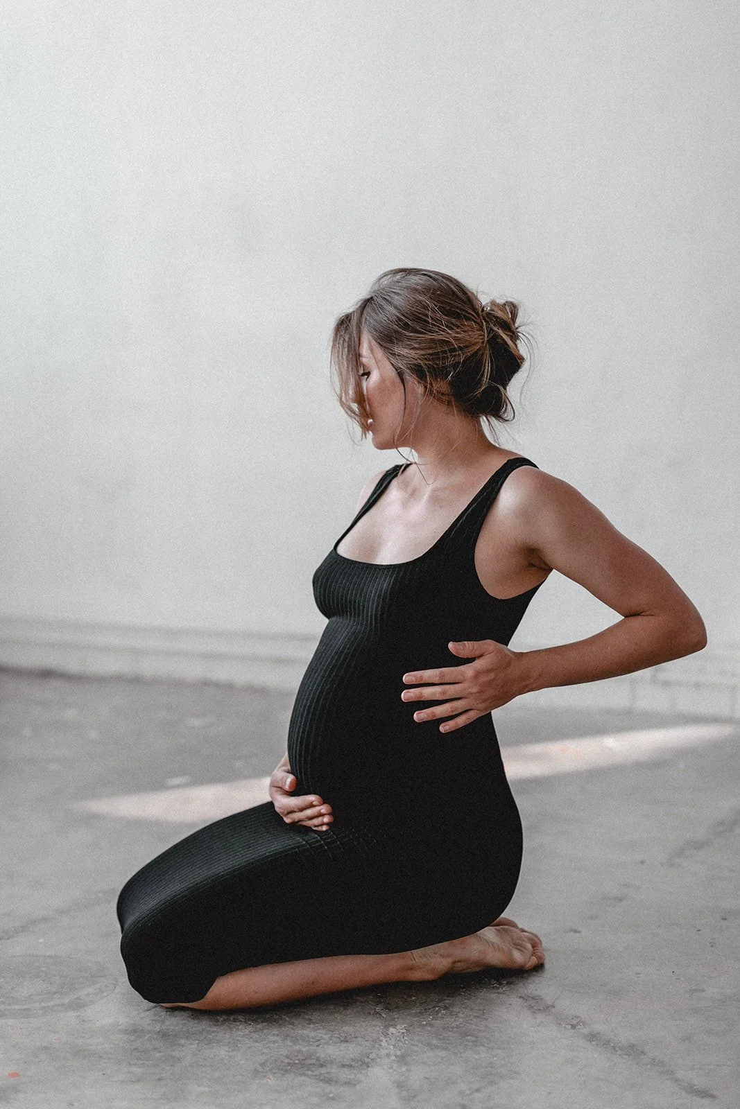 Schwangere Frau in Schwarz in Meditationshaltung auf Knien, hält ihren Babybauch, Auge geschlossen, in einem neutralen Raum.