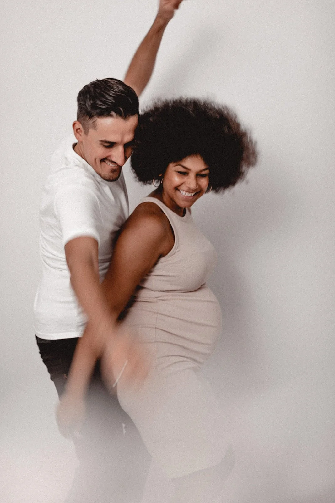 Ein glückliches Paar, das zusammen tanzt, die schwangere Frau trägt ein beige Kleid, der Mann trägt ein weißes T-Shirt, beide lachen und bewegen sich im Takt.