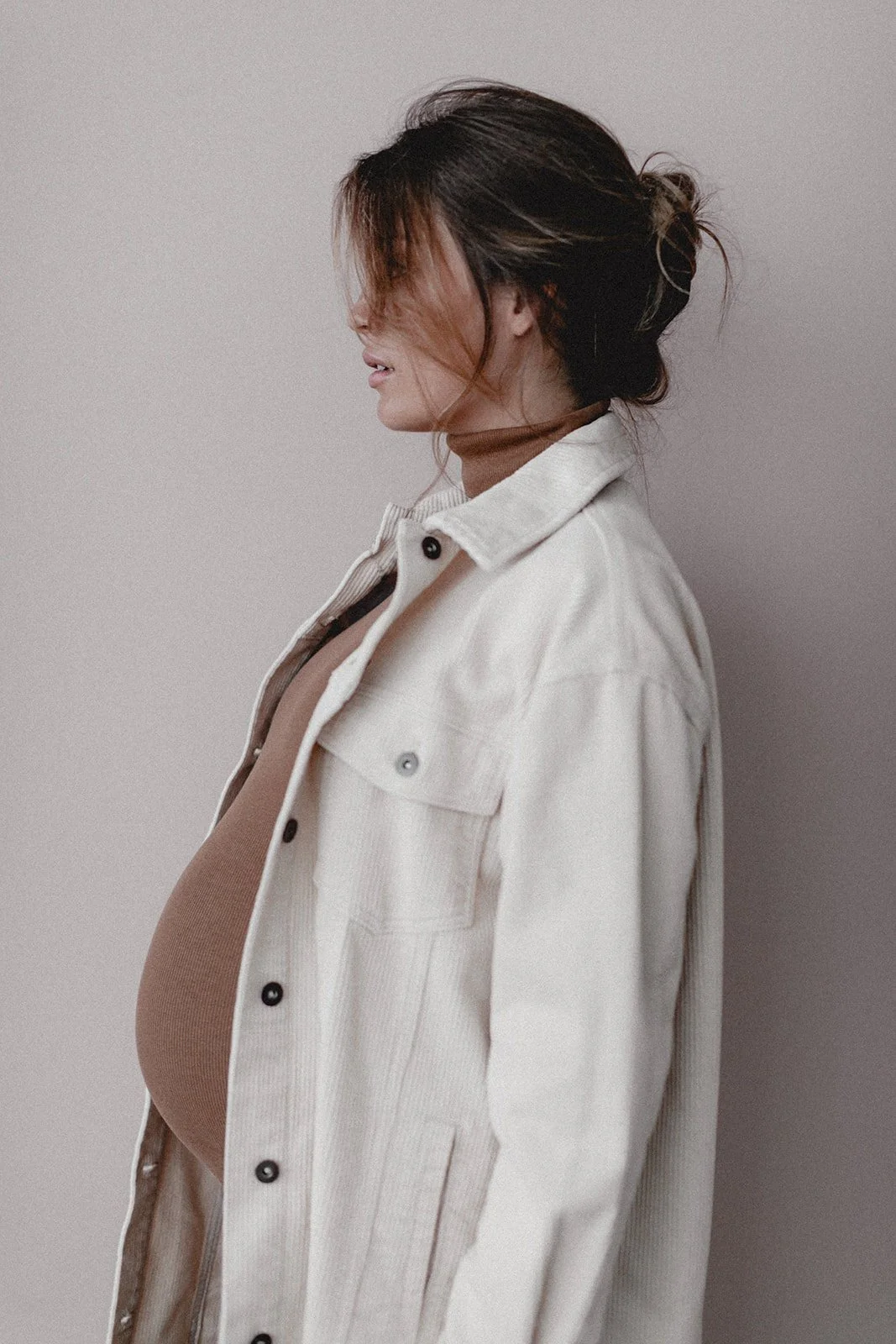 Eine schwangere Frau trägt ein braunes Oberteil und einen weißen Kittel, steht seitlich vor einer grauen Wand.