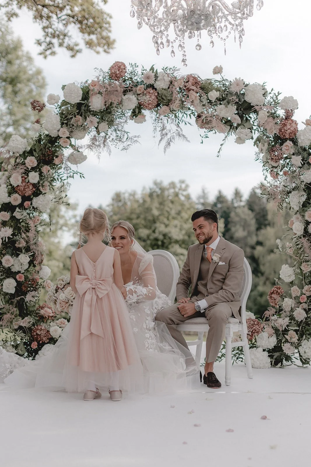 Hochzeitsszene mit einem Brautpaar, einem Kind, einer Blumensäule und einer Kristallleuchterdecke im Freien.