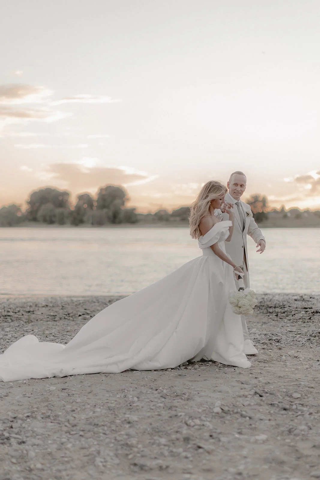 Ein Brautpaar am Ufer bei Sonnenuntergang, die Braut trägt ein weißes Hochzeitskleid mit Schleppe und hält einen Blumenstrauß, der Bräutigam trägt einen hellen Anzug, im Hintergrund Wasser und Bäume.