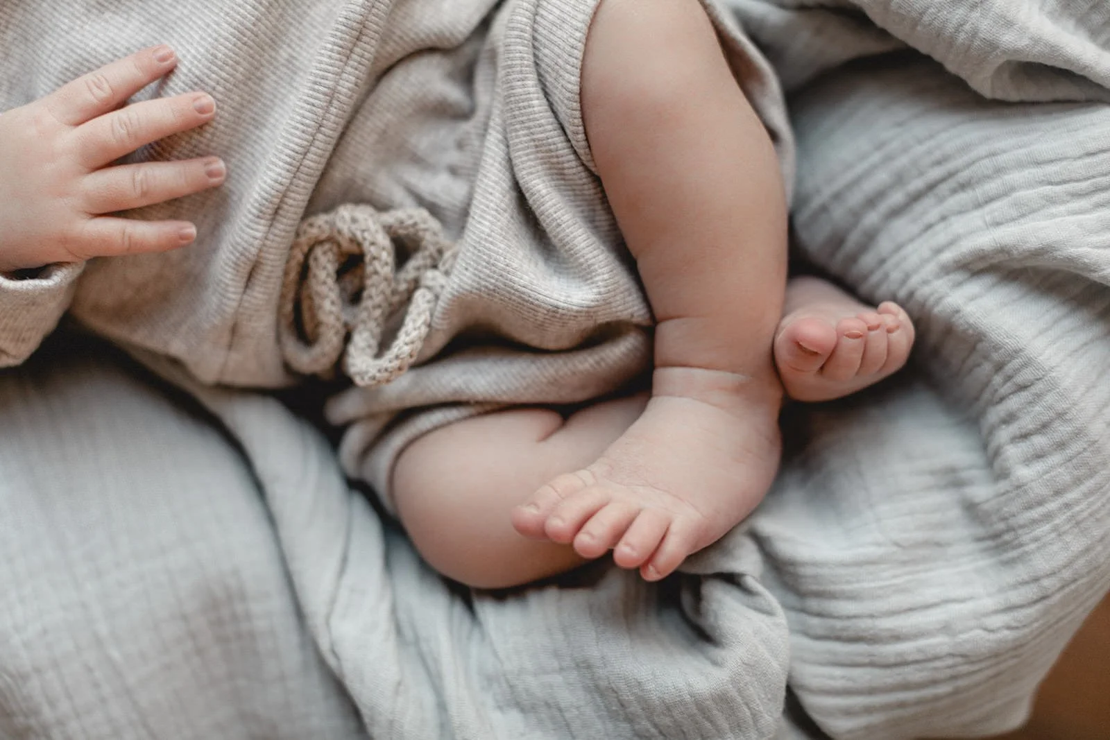 Nahaufnahme eines Babys, das in neutralen, hellgrauen Stoffen liegt und seine kleinen Hände und Füße zeigt.