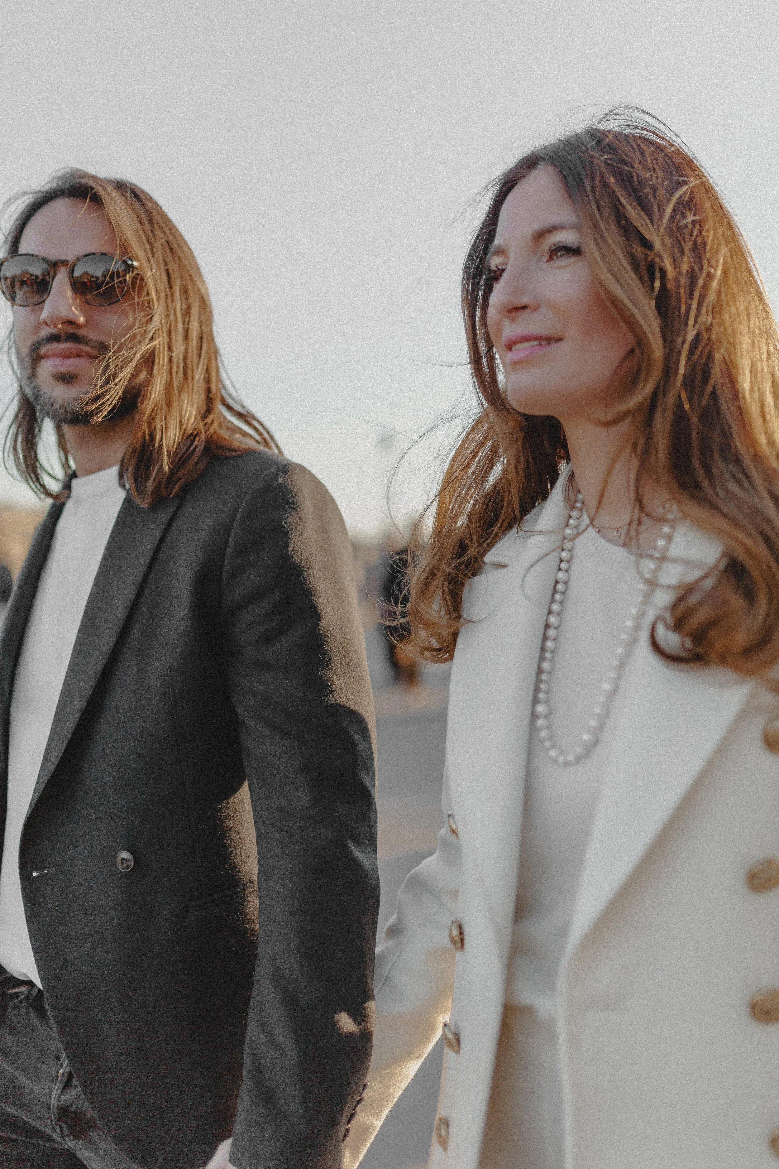 Ein Mann und eine Frau, beide in eleganten Outfits, gehen im Freien bei Sonnenuntergang. Der Mann trägt eine schwarze Jacke, die an den Schultern mit einem einzigartigen Farbverlauf verziert ist, und Sonnenbrillen. Die Frau trägt einen cremefarbenen 