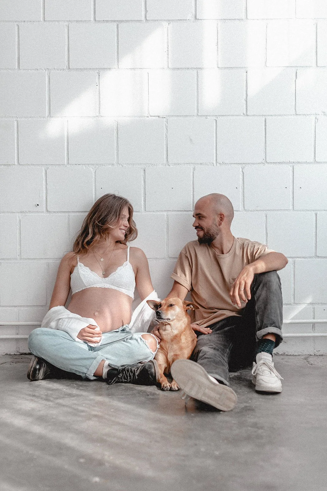 Ein Paar sitzt auf dem Boden vor einer weißen Ziegelwand, mit einem Hund zwischen ihnen, sie lächeln sich an.