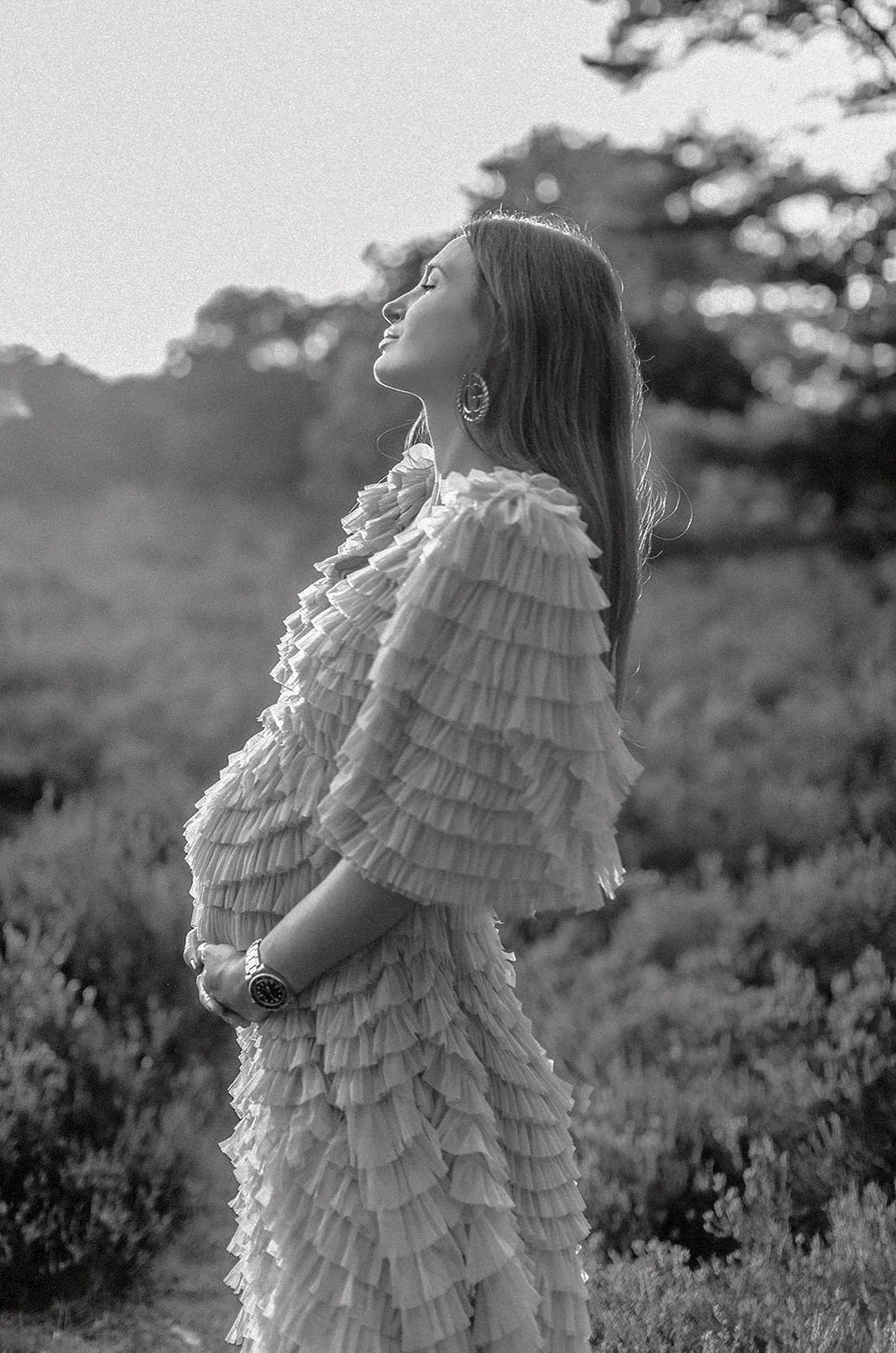 Eine Frau in einem langen, mehrlagigen, gerüschten Kleid steht im Freien und genießt die Sonne, mit Bäumen im Hintergrund, in schwarz-weiß.