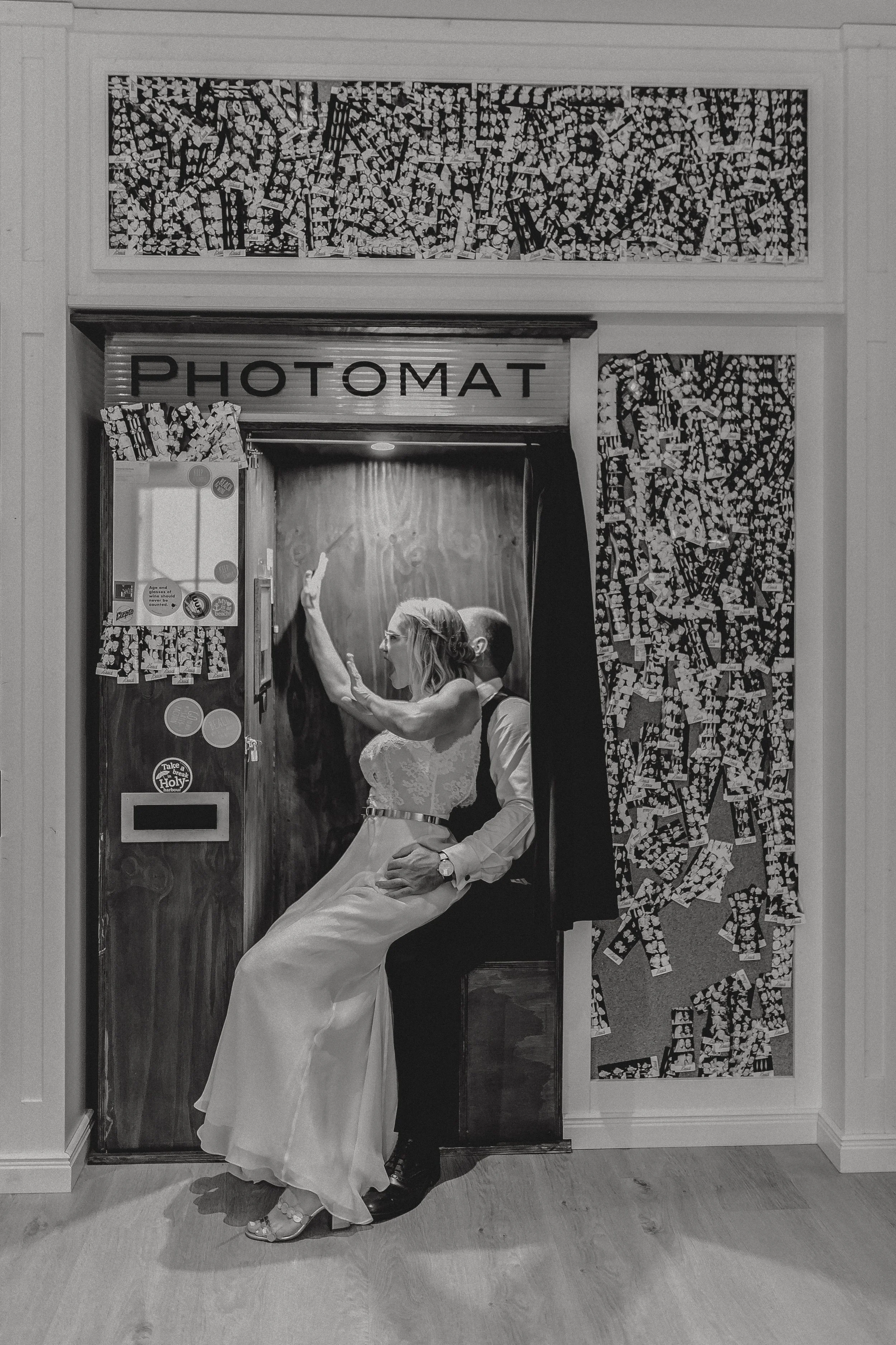 Ein Brautpaar in einem Fotokabine, die Braut trägt ein weißes Hochzeitskleid, der Bräutigam trägt einen Anzug. Sie posieren für ein Foto, das im Mittelpunkt steht. Die Fotokabine ist mit dem Schild 'PHOTOMAT' oberhalb gekennzeichnet, und die Umgebung
