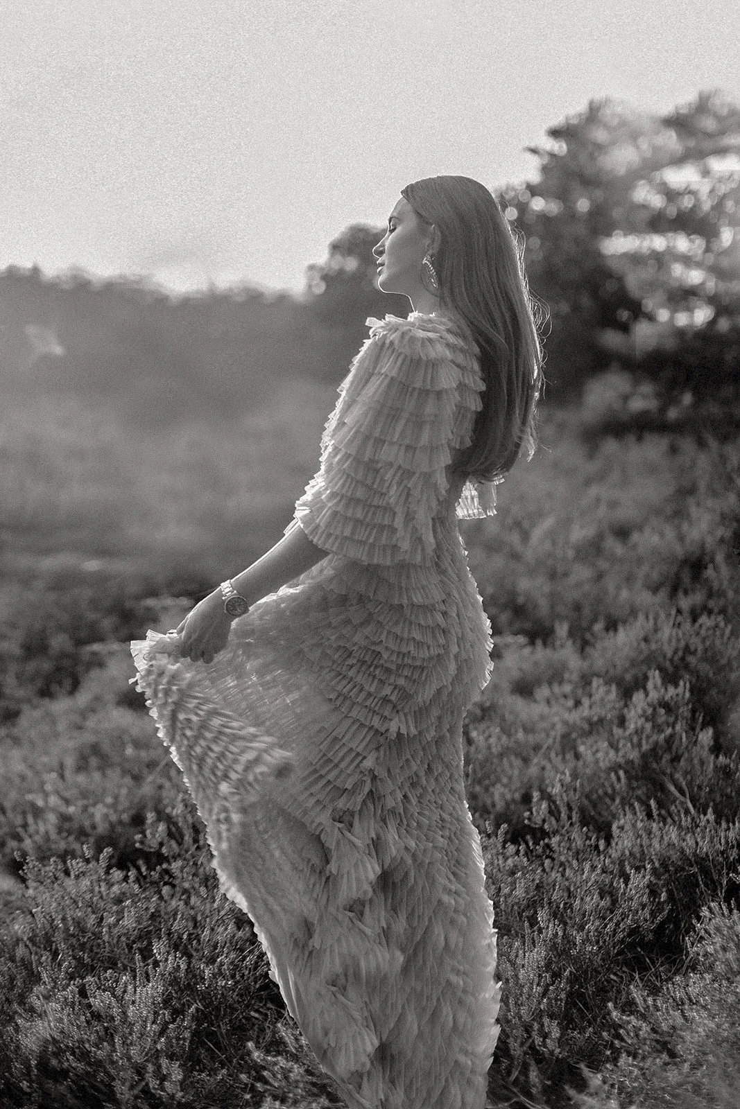 Frau in einem langen, mehrlagigen, Rüschen kleid steht im Freien bei Sonnenuntergang, blickt nach rechts, umgeben von Büschen und Bäumen.