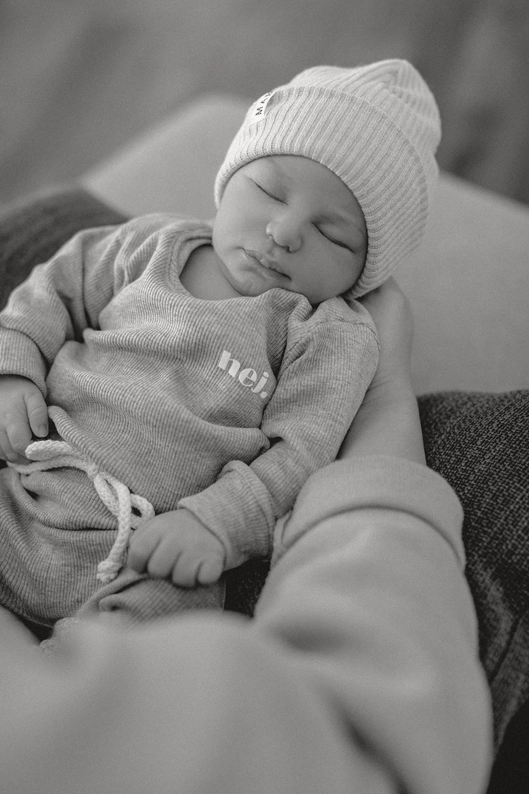 Schlafendes Baby mit Mütze, getragen in einem Arm, in Schwarz-Weiß-Fotografie.
