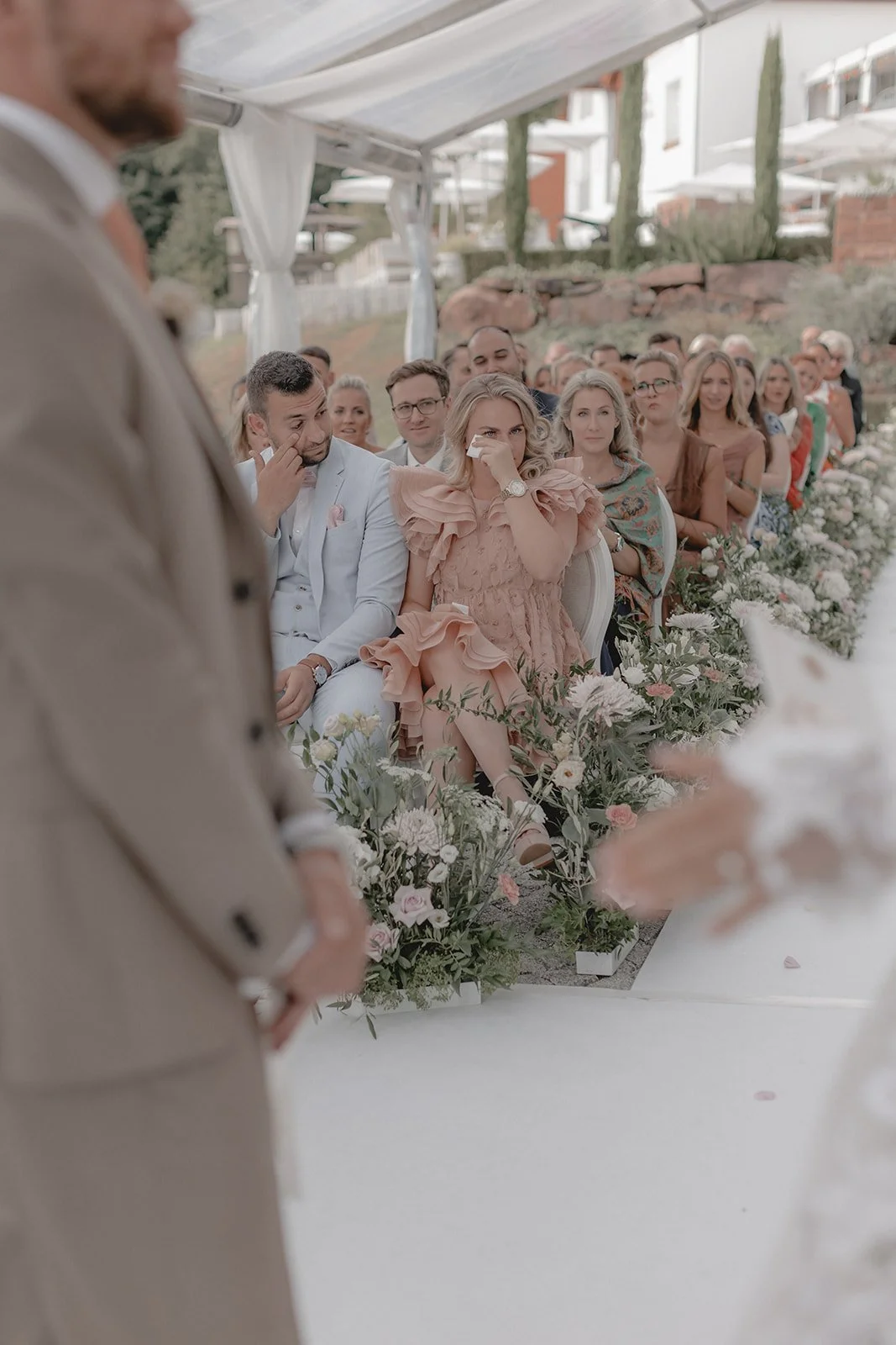 Menschen bei einer Hochzeitszeremonie, sitzend unter einer transparenten Überdachung, mit vielen Blumen und Gräsern im Hintergrund.