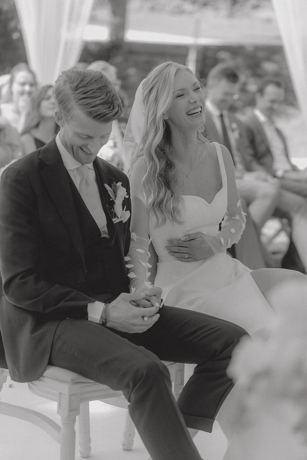 Braut und Bräutigam sitzen bei ihrer Hochzeit, lachen und halten Händchen. Im Hintergrund sitzen Gäste in einem Zelt.