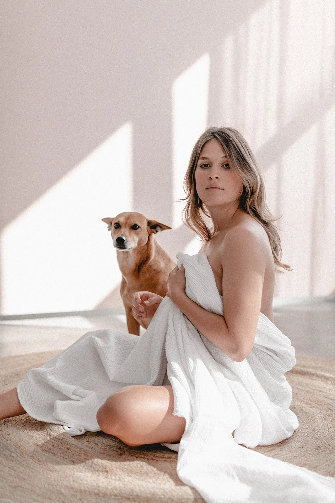 Junge Frau sitzt nackt auf einem Teppich, hält ein weißes Handtuch vor ihrer Brust, mit einem Hund im Hintergrund, in einem Raum mit Sonnenlicht und Schatten an der Wand.