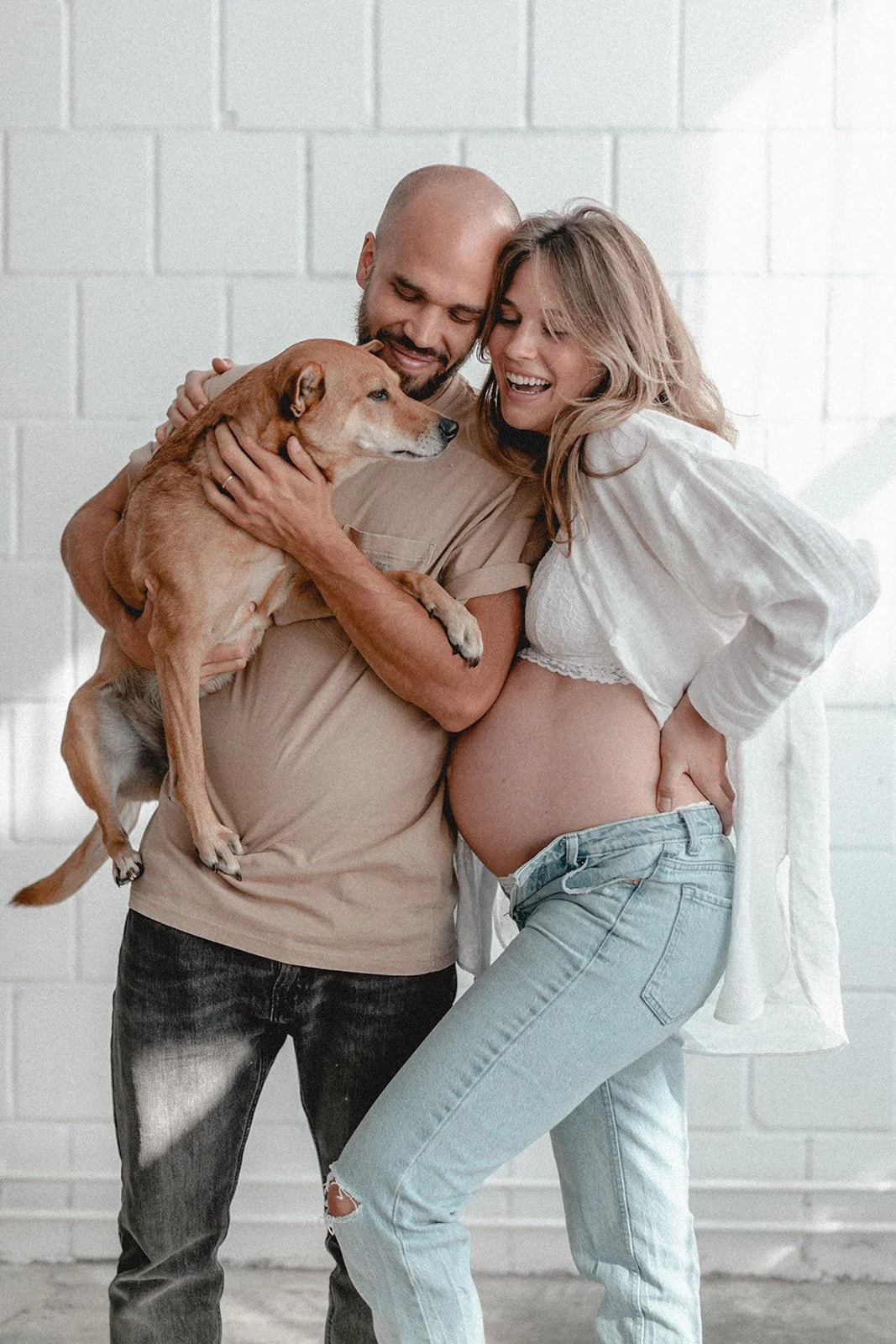 Ein Paar freut sich, während die schwangere Frau ihren Babybauch zeigt, begleitet von einem Mann und einem Hund in einem hellen Raum mit weißen Ziegelwänden.