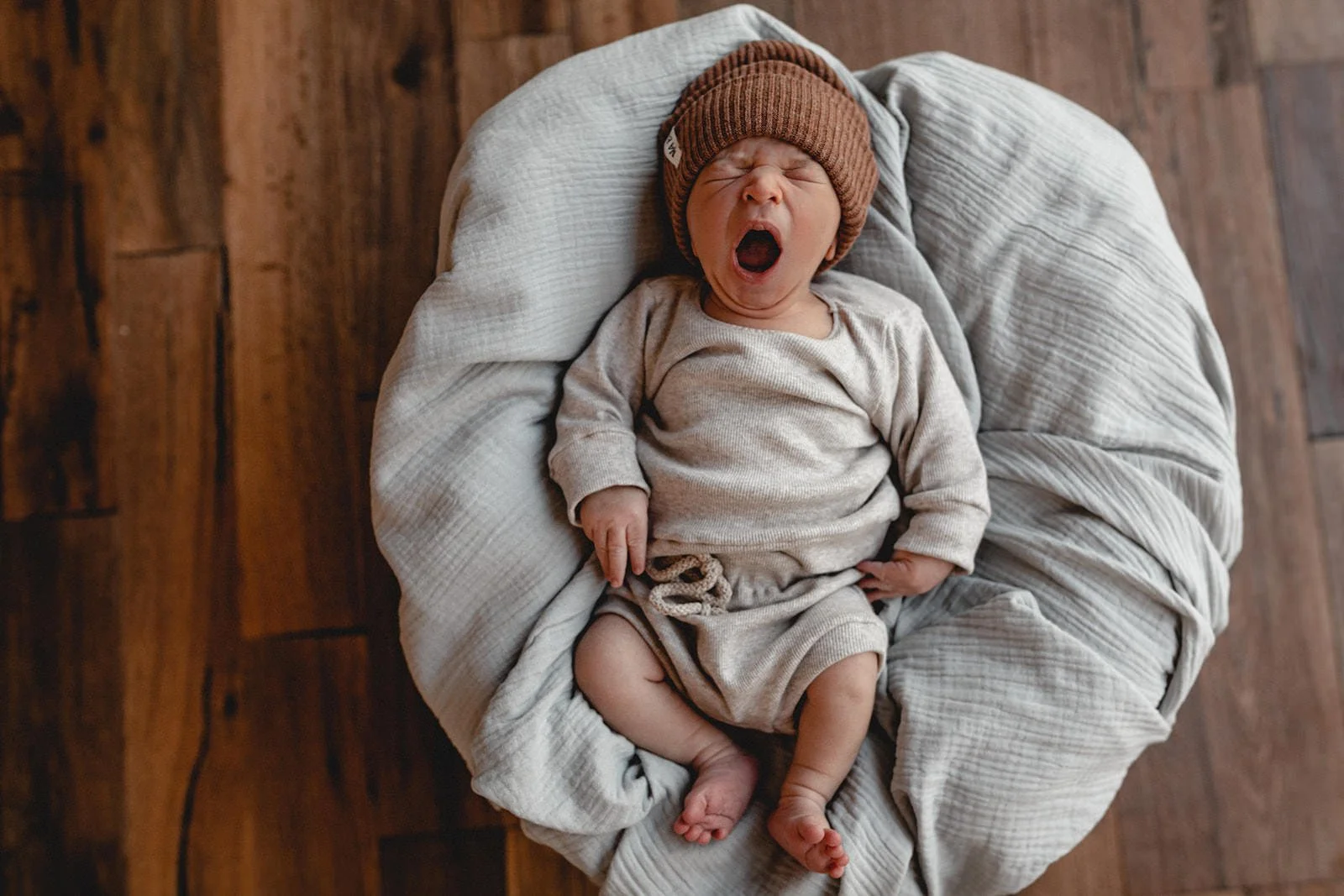 Ein schreiendes Baby liegt auf einem dicken, hellgrauen Kissen auf einem Holzboden. Das Baby trägt eine braune Mütze und bequeme Kleidung in hellen, neutralen Tönen.
