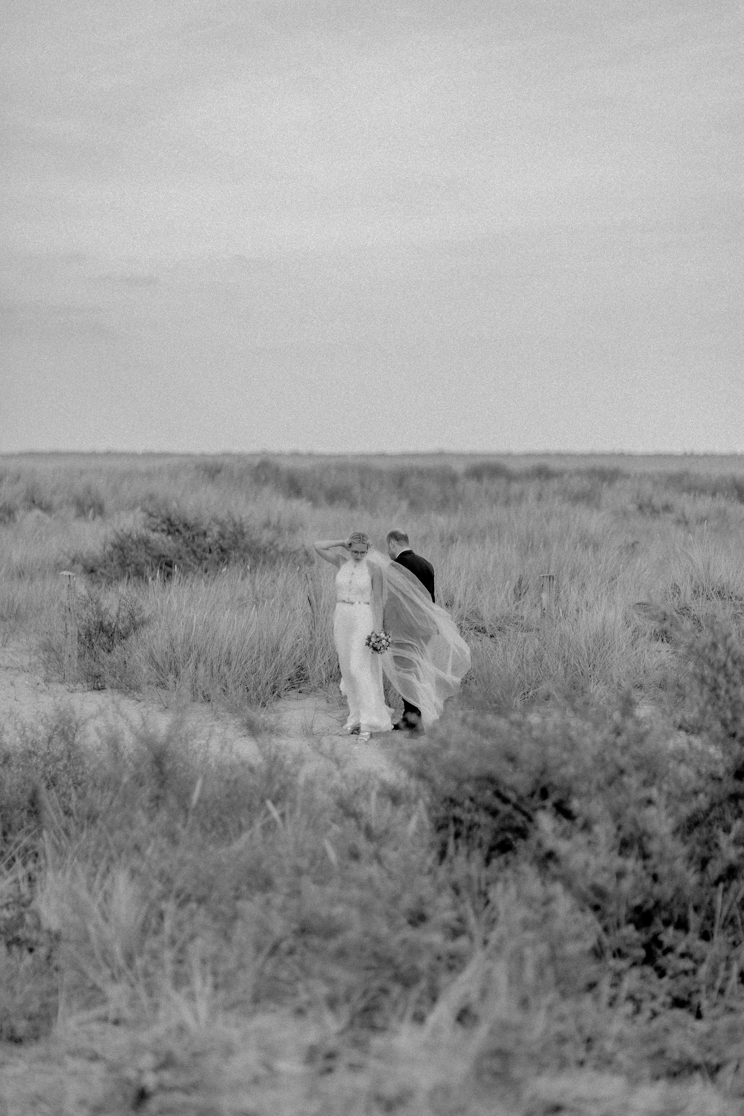 Zwei Menschen in formeller Kleidung, eines mit einem Blumenstrauß, gehen durch eine Graslandschaft.