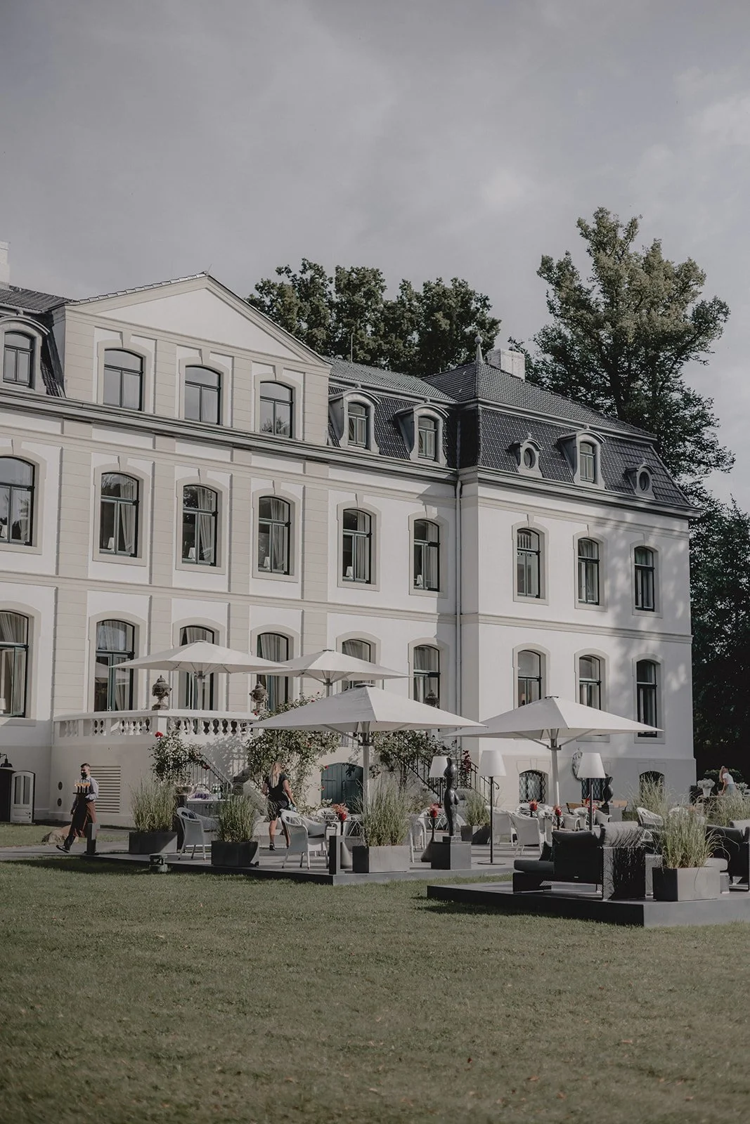 Schloss Weissenhaus. Ein großes, weißes, mehrstöckiges Herrenhaus mit zahlreichen Fenstern und einem Mansarddach. Im Vordergrund befindet sich ein Garten mit mehreren Tischen und Sesseln, sowie einigen Personen, die den Garten nutzen.