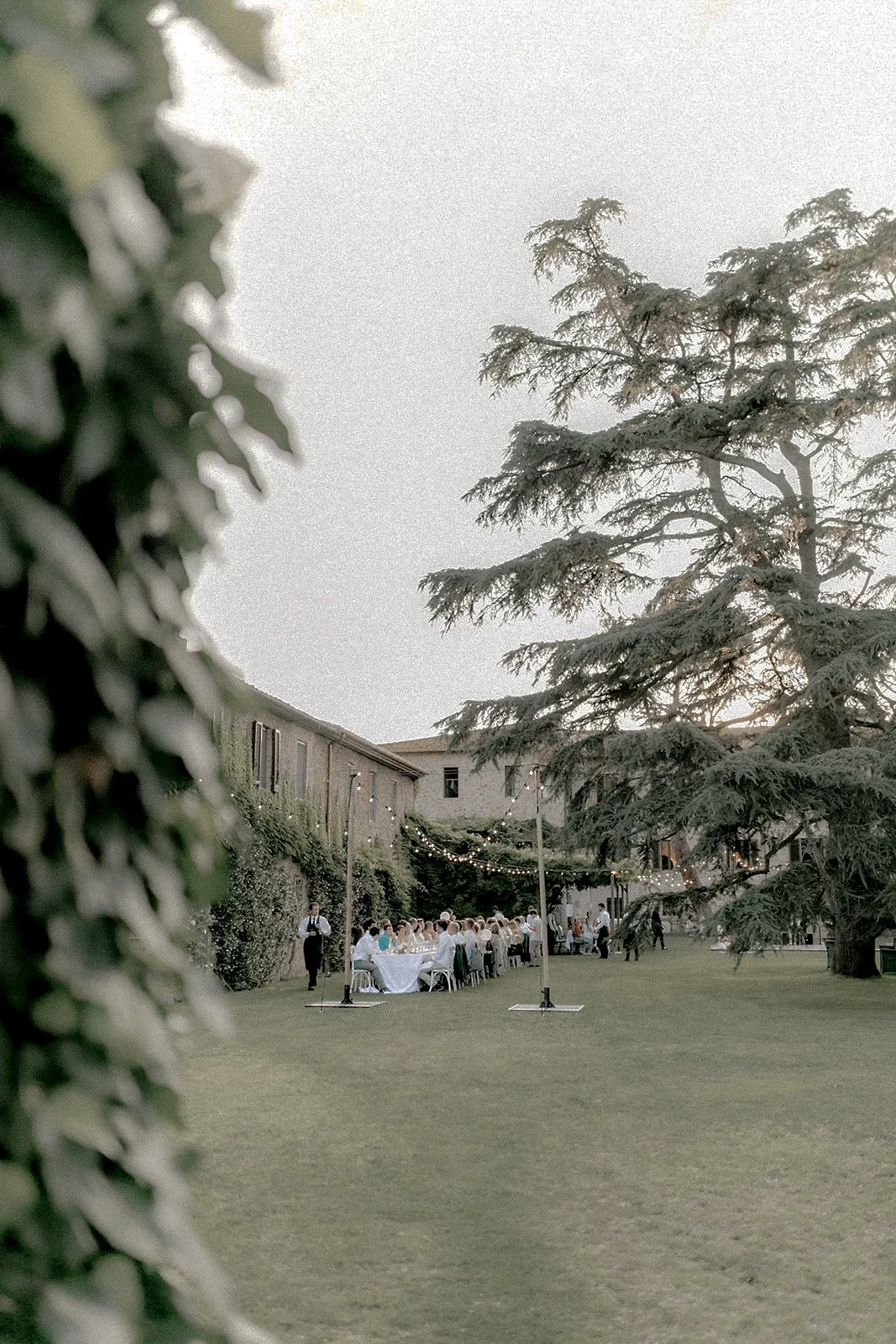 Eine Outdoor-Gaststätte bei Sonnenuntergang mit Tischen, Stühlen, festlicher Beleuchtung und Gästen, umgeben von Bäumen und Gebäuden.