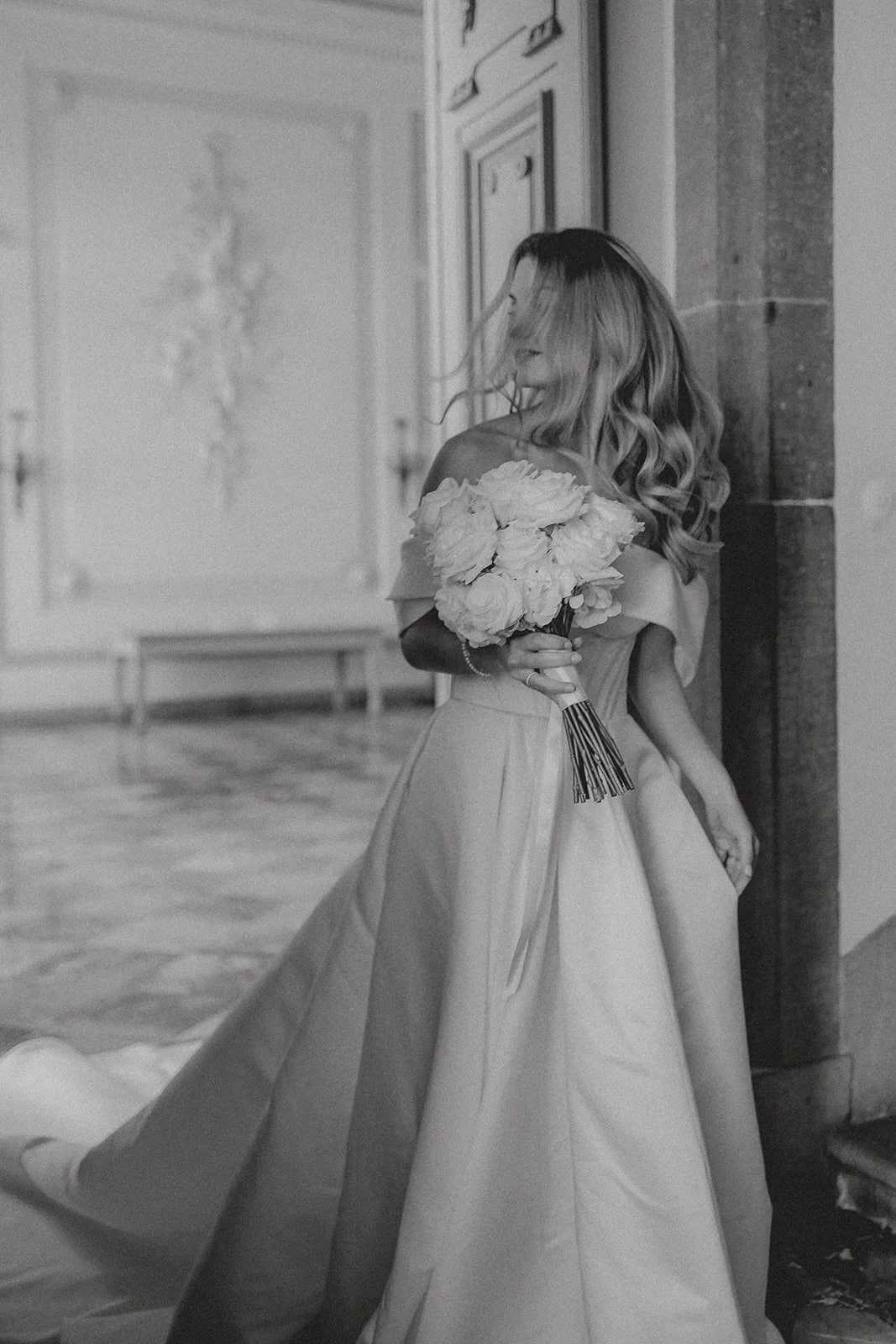 Eine Frau in Hochzeitskleidung hält einen Bouquet aus großen Blumen und steht in einem Raum mit antiken Möbeln.