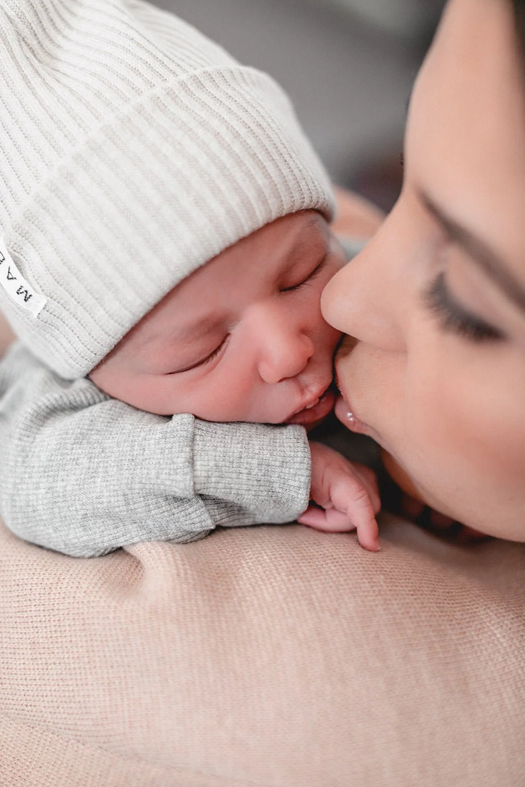 Nahaufnahme einer Mutter, die ihr neugeborenes Baby liebevoll küsst. Das Baby trägt einen hellgrauen Strickmütze und befindet sich im Babybody auf der Schulter der Mutter.