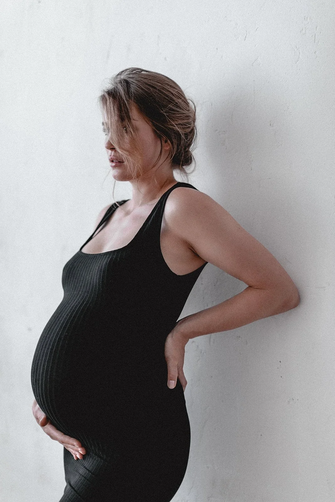 Schwangere Frau in schwarzem Kleid lehnt an weißer Wand, Blick nach unten, mit Hand auf Babybauch.