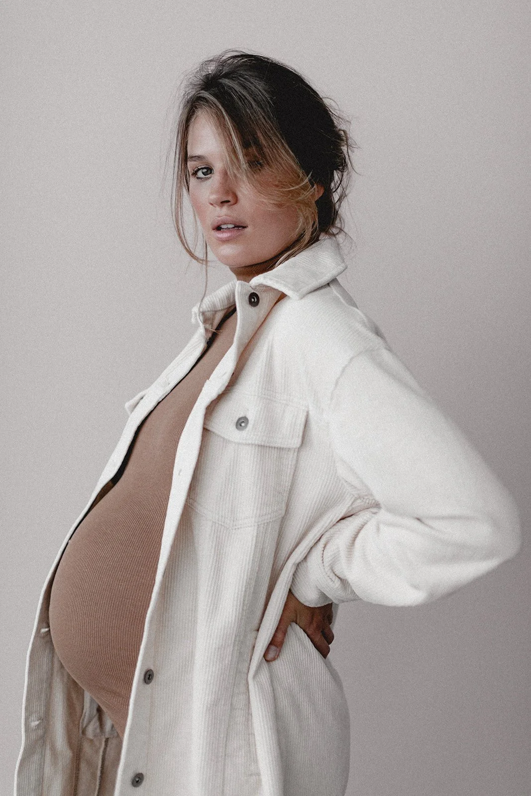 Eine schwangere Frau trägt ein offenes cremefarbenes Jacke und einen beigen Oberteil, steht seitlich vor einem neutralen grauen Hintergrund.