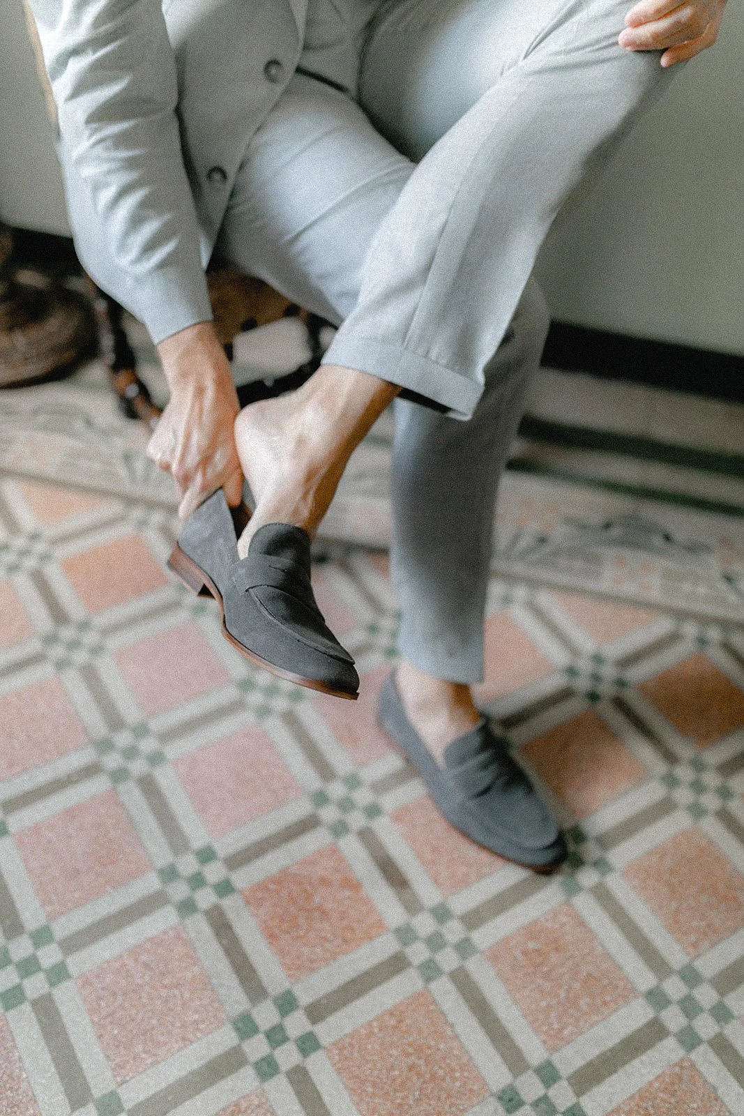 Eine Person in grauem Anzug setzt sich auf einen Stuhl, während sie einen schwarzen Lederschuh anzieht, auf einem Fuß steht ein weiteres schwarzes Schuhpaar, die Person sitzt auf einem gepflasterten Boden.