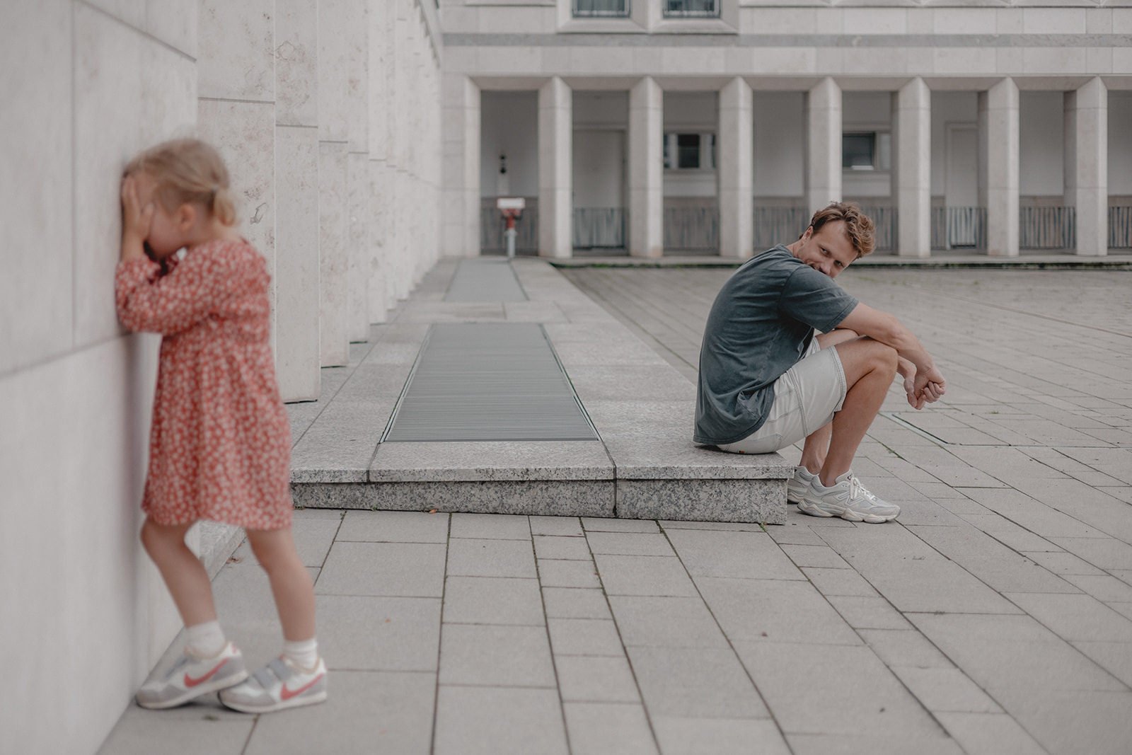 Ein Mann sitzt auf einer steinernen Plattform und schaut skeptisch auf eine kleine, verängstigte Mädchen, die an einer Mauer steht und sich die Ohren zuhält.