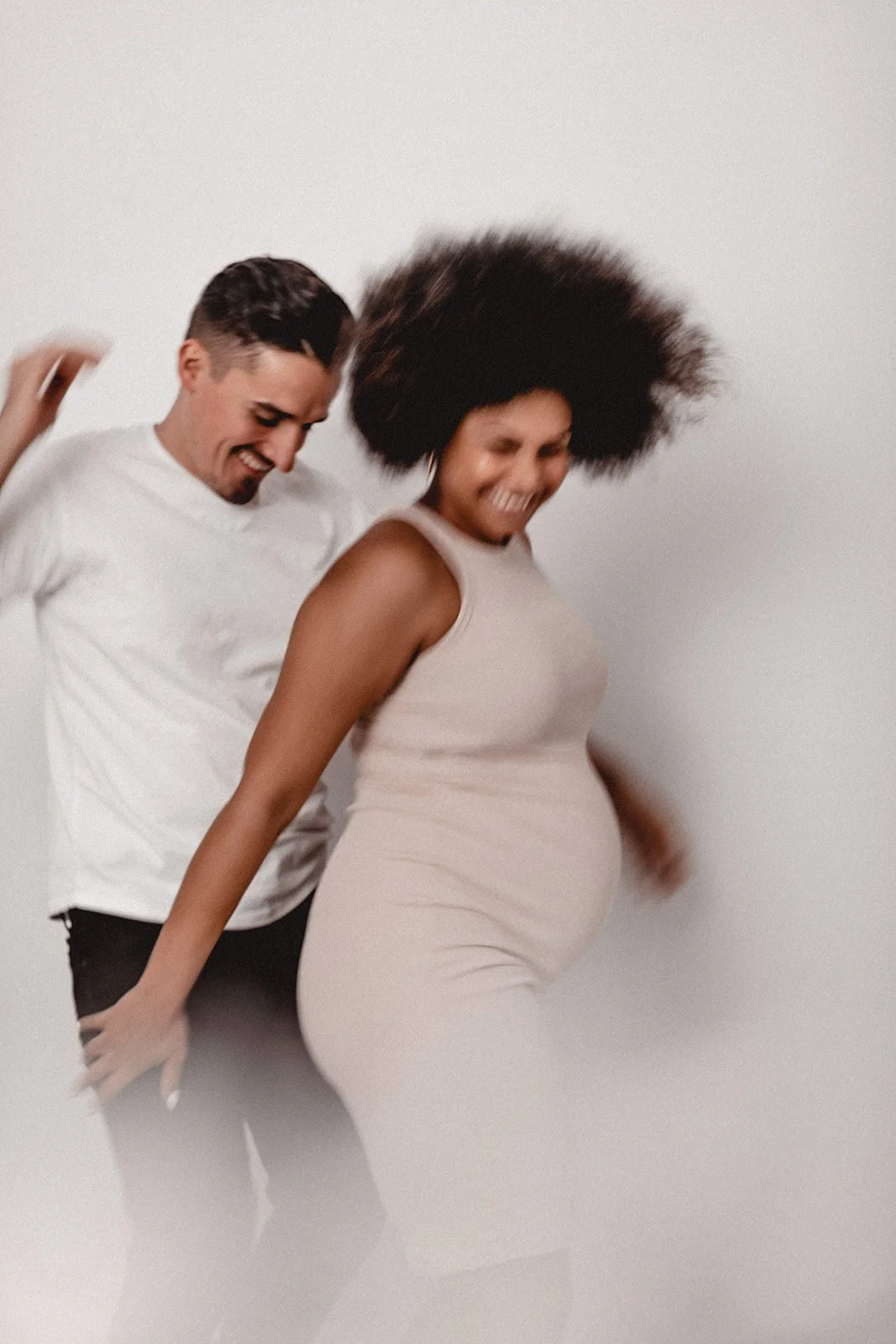 Ein lachendes Paar tanzt zusammen, der Mann trägt ein weißes T-Shirt, die schwangere Frau trägt ein beiges Kleid.