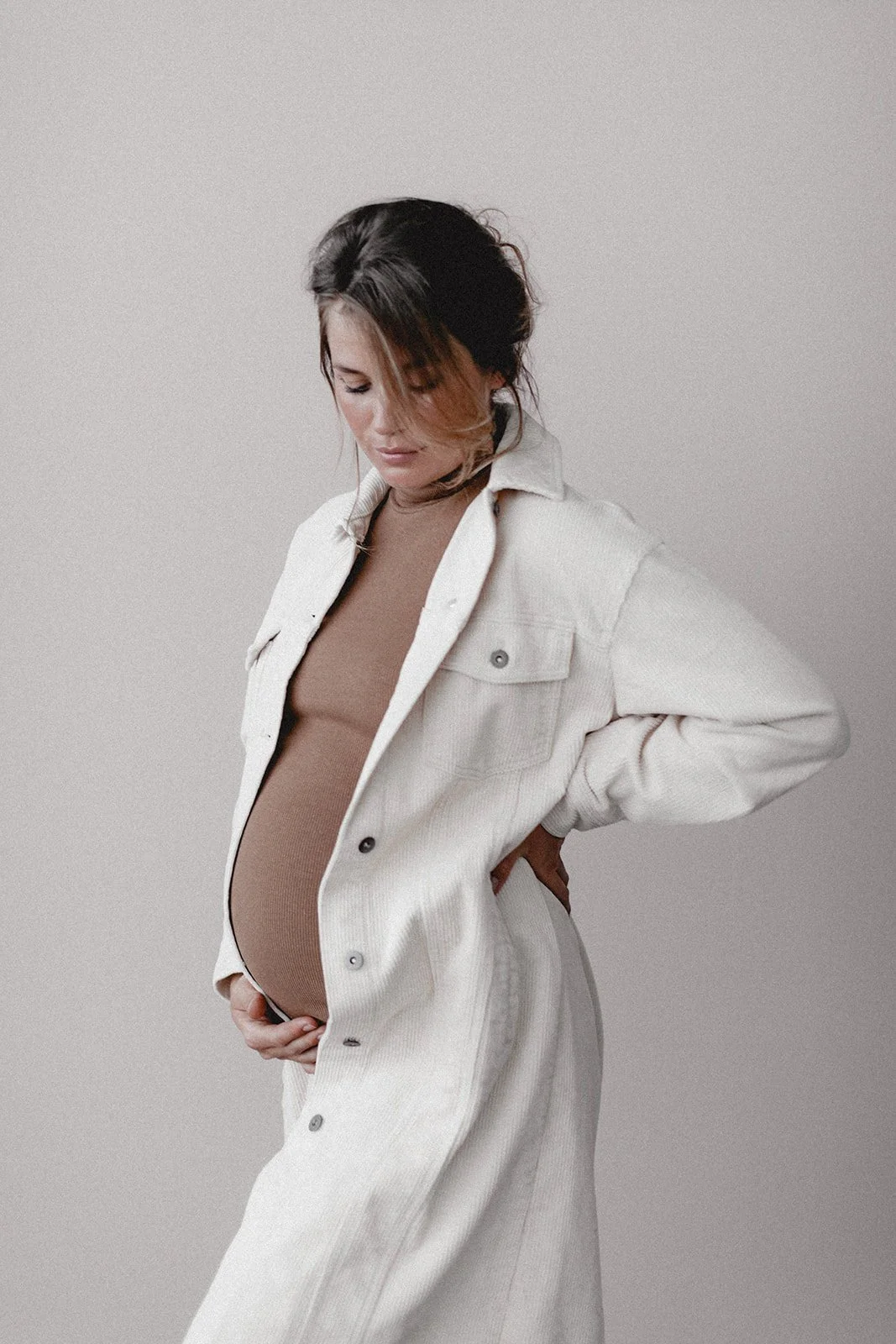 Eine schwangere Frau mit dunklen, teilweise gelblich gefärbten Haaren trägt einen beigen Body und eine weiße Jeansjacke, und steht vor einem grauen Hintergrund.