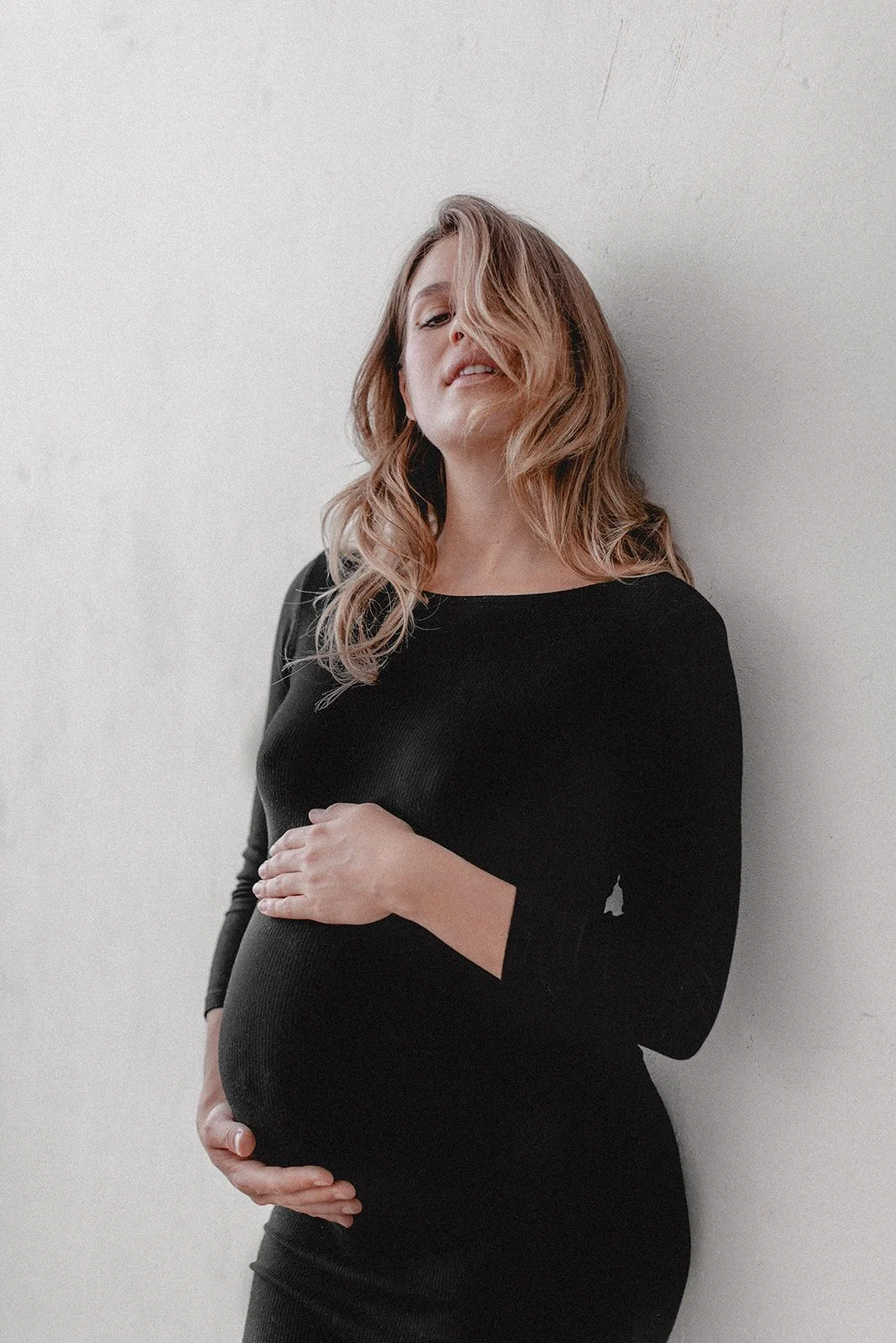 Eine schwangere Frau in einem schwarzen Kleid lehnt an einer weißen Wand und hält ihre Babybauch.