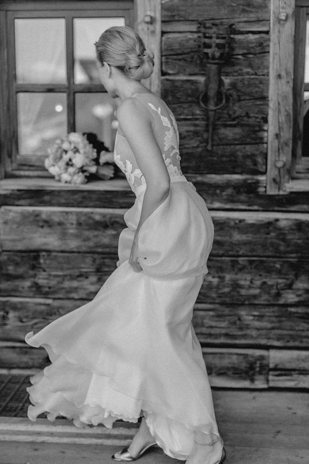 Eine Frau in einem weißen, eleganten Kleid steht in einem rustikalen Raum, Blick nach unten, mit einem Brautstrauß auf einer Fensterbank im Hintergrund, im Schwarz-Weiß-Format.