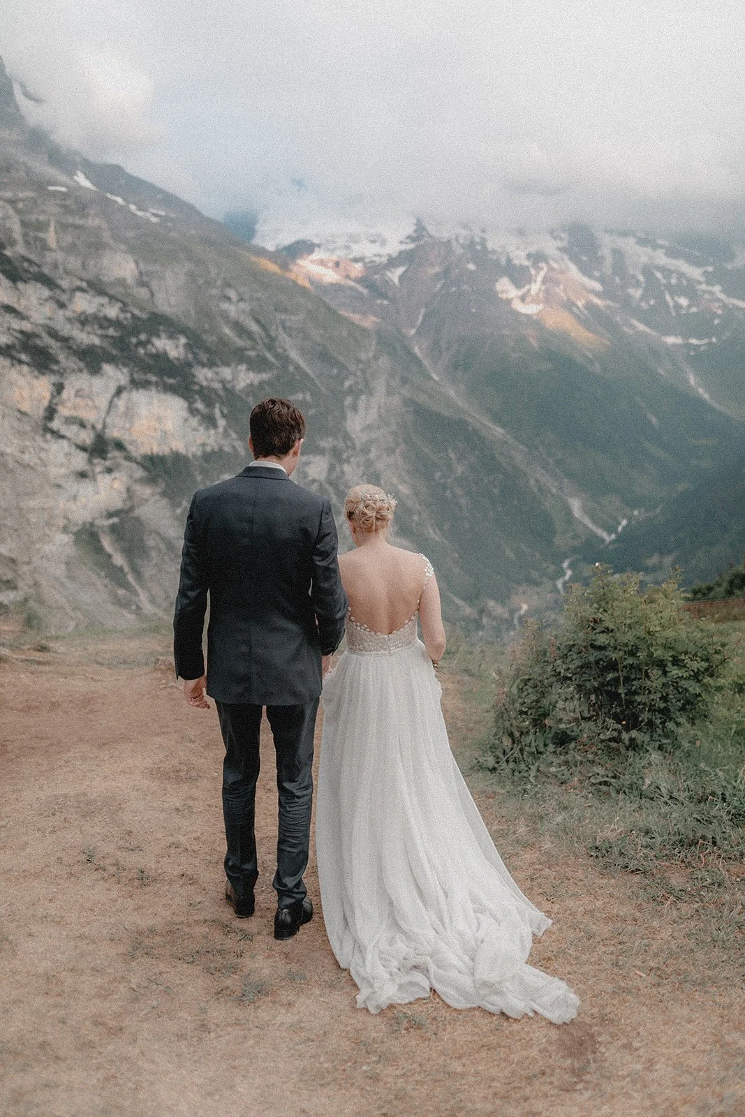 Ein Paar in Hochzeitskleidung geht auf einer Wanderung in den Bergen, mit Blick auf schneebedeckte Gipfel und eine bewölkte Himmelspanne.