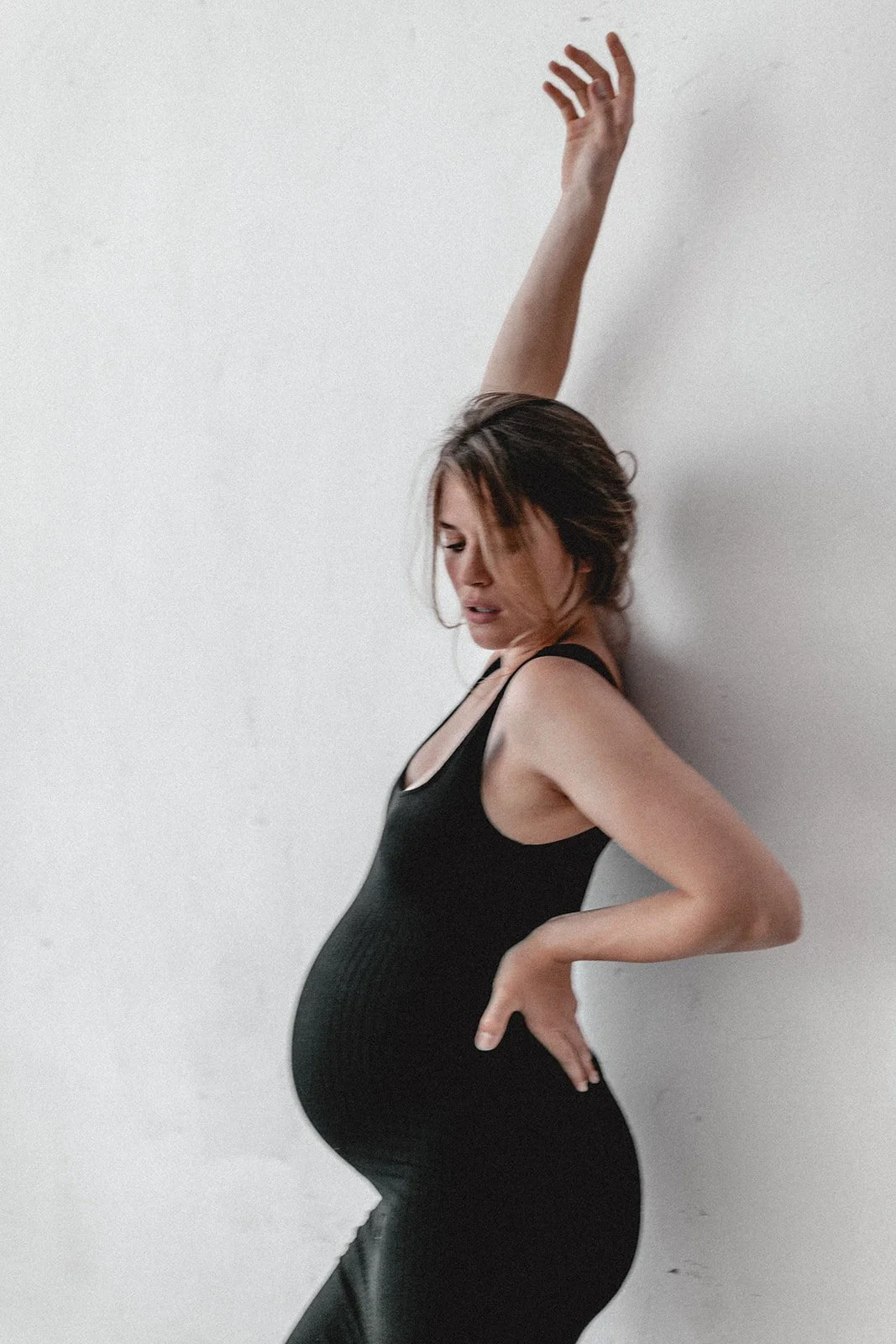 Schwangere Frau in schwarzem Kleid lehnt an weißer Wand, mit Arm nach oben und Hand auf Hüfte.
