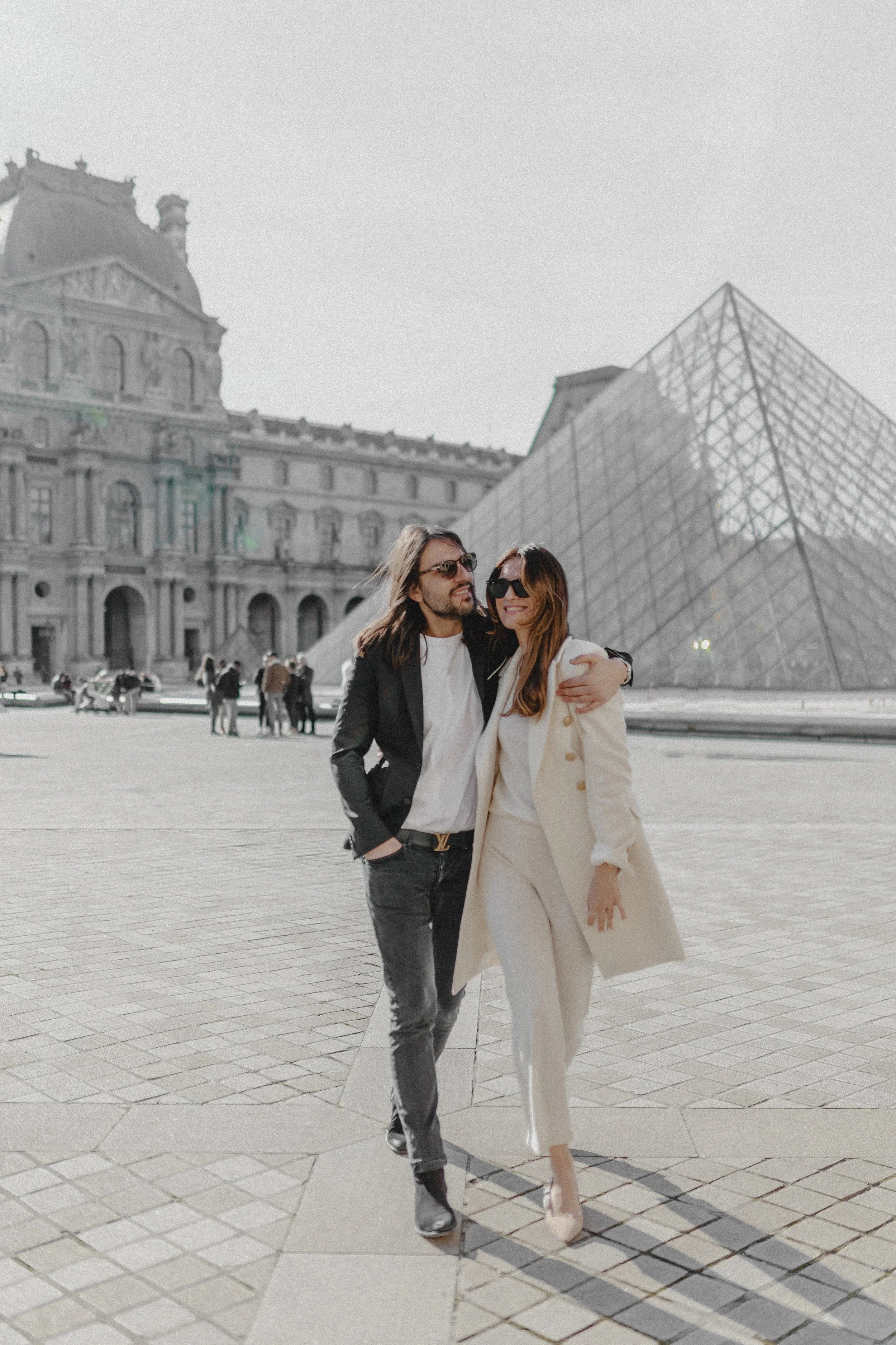 Ein Paar in modischer Kleidung, lächelnd, umarmt sich vor dem Louvre in Paris, Frankreich, bei sonnigem Wetter.