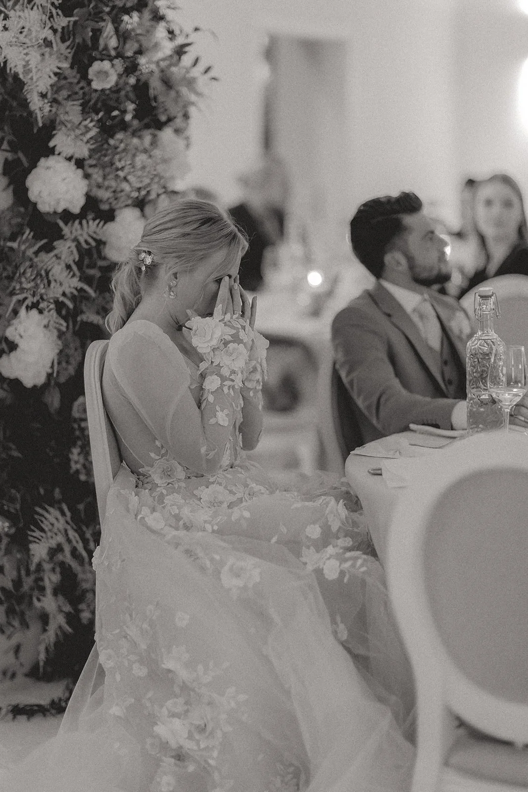 Eine weinende Frau in einem eleganten, langärmligen, floral bestickten Kleid sitzt an einem gedeckten Tisch bei einer Feier. Im Hintergrund sitzen weitere Gäste, darunter ein Mann in Anzug und eine Frau, die im Gespräch sind. Eine große Blumenpracht 