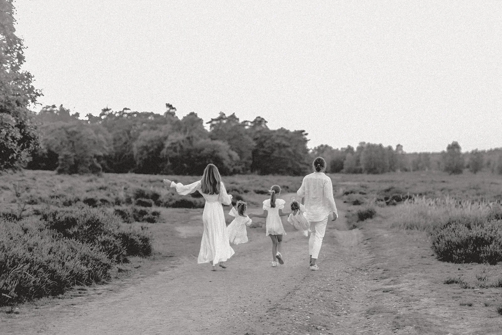 Eine Familie, bestehend aus zwei Erwachsenen und vier Kindern, läuft auf einem offenen Feld in Schwarz-Weiß. Die Erwachsenen tragen weiße Kleidung, die Kinder tragen Kleider. Die Familie bewegt sich in Richtung des Waldes am Horizont.