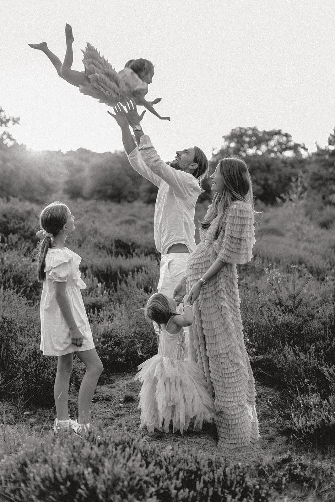 Eine Familie im Freien, der Vater hebt ein kleines Mädchen, das ein Flügelkostüm trägt, hoch in die Luft, während die Mutter und zwei ältere Kinder zuschauen. Es ist ein fröhliches Spiel im Freien bei Sonnenuntergang.