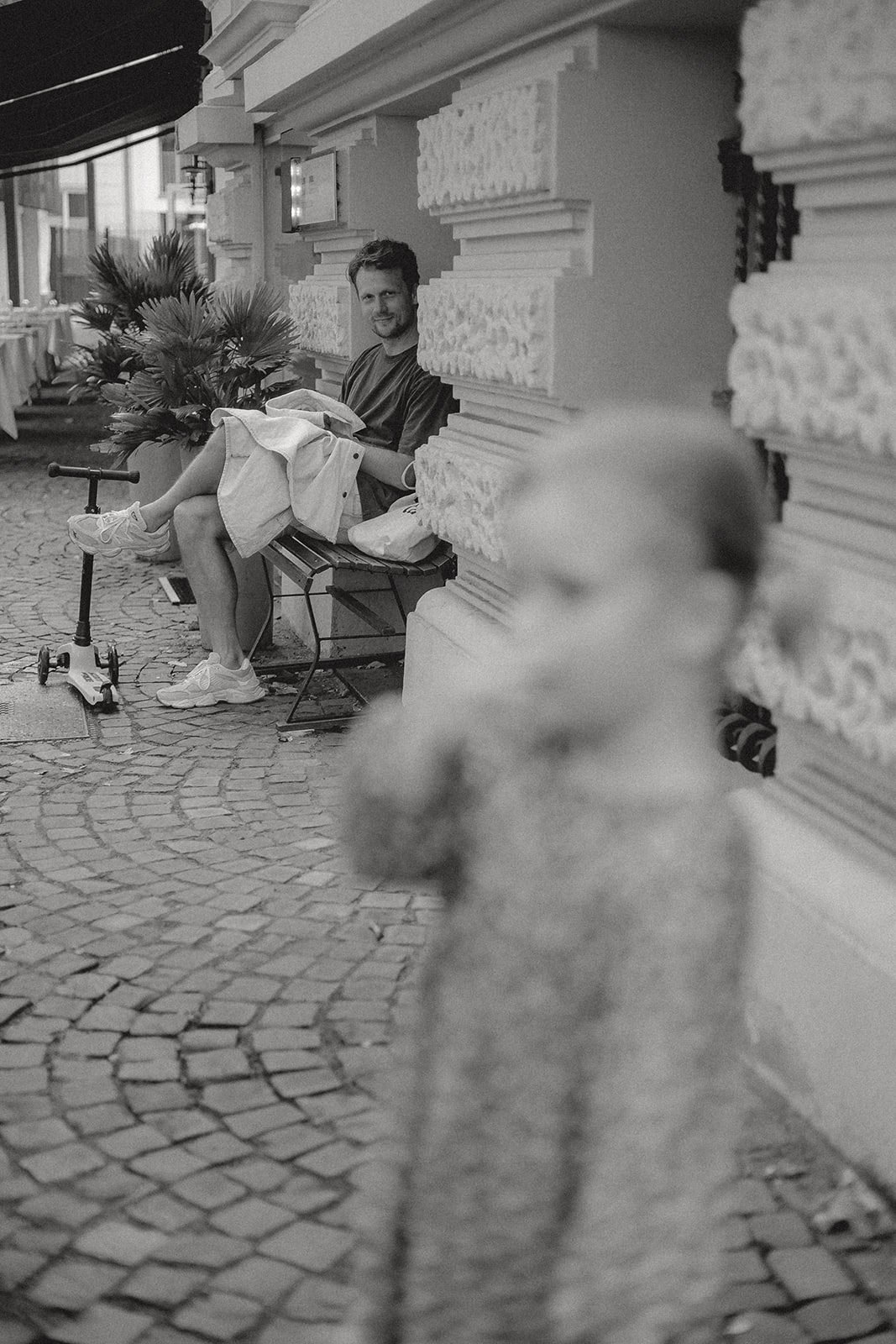 Ein Mann sitzt auf einer Bank auf der Straße neben einem kleinen Hund. Im Vordergrund ist ein verschwommener Hund zu sehen, im Hintergrund eine Blume und ein Kind mit einer Piaggio-Kleinstadt.