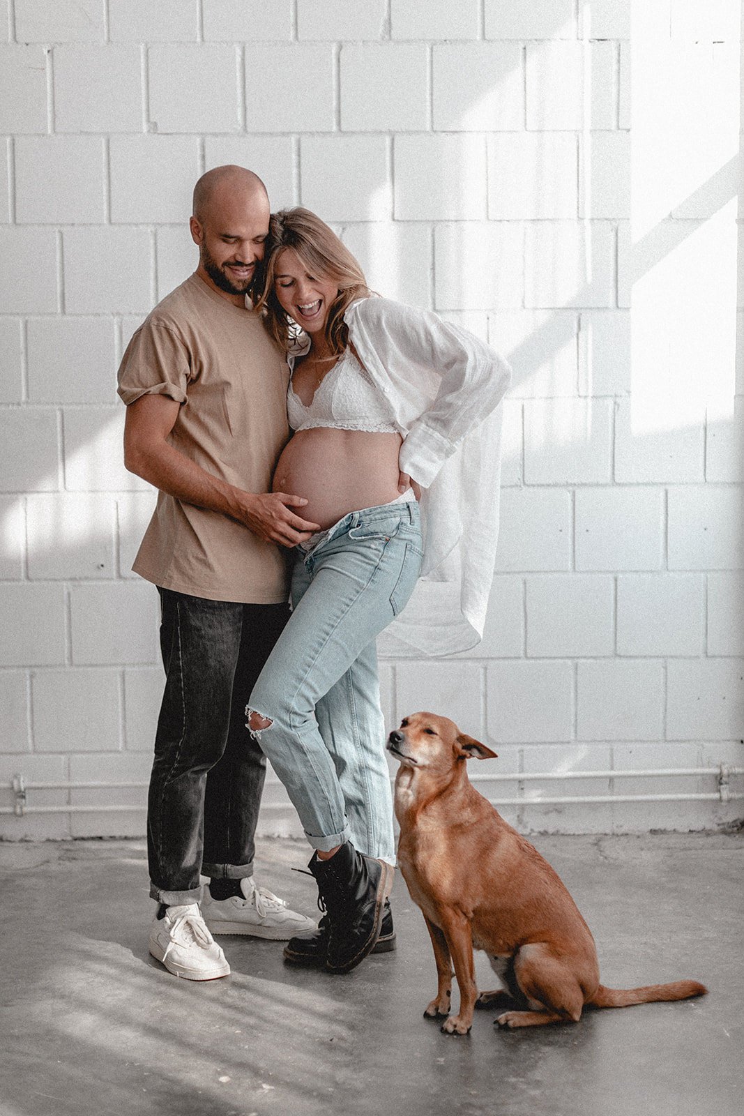 Ein werdendes Elternpaar steht lachend vor einer weißen Wand, sie streicht auf den Bauch der Frau, während ein Hund bequem neben ihnen sitzt und schaut.
