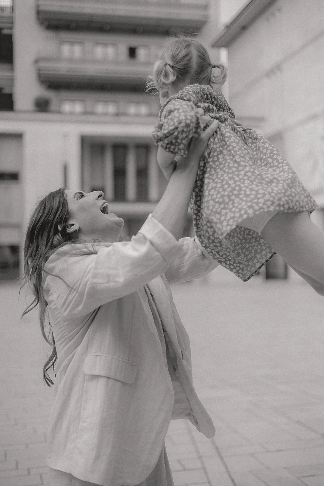 Zwei Frauen spielen mit einem Kind auf einem Balkon, die eine Frau hält das Kind hoch, beide lachen, im Hintergrund sind moderne Wohngebäude sichtbar.
