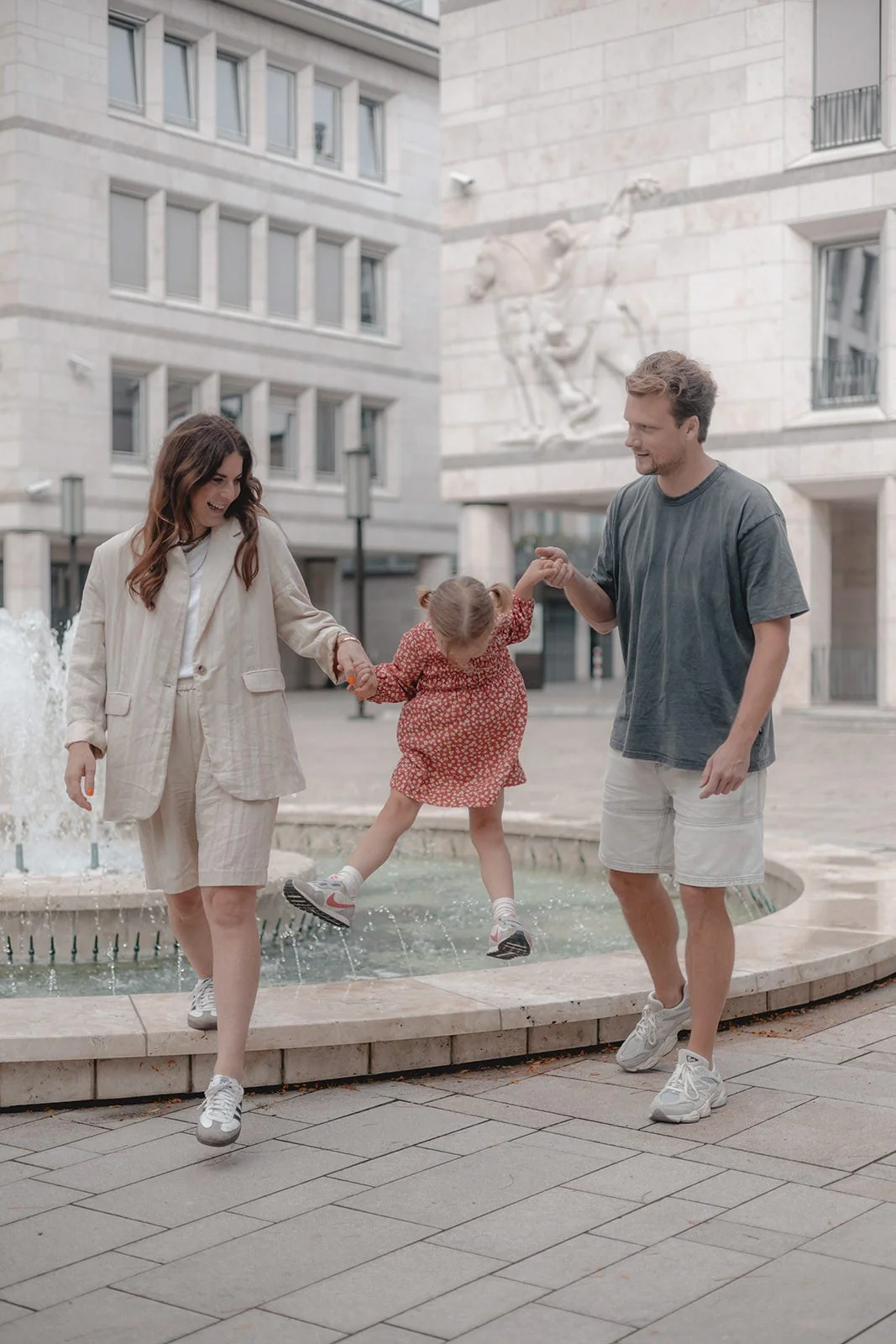 Familie beim Spielen an einem Brunnen in einem urbanen Bereich mit modernen Gebäuden im Hintergrund.