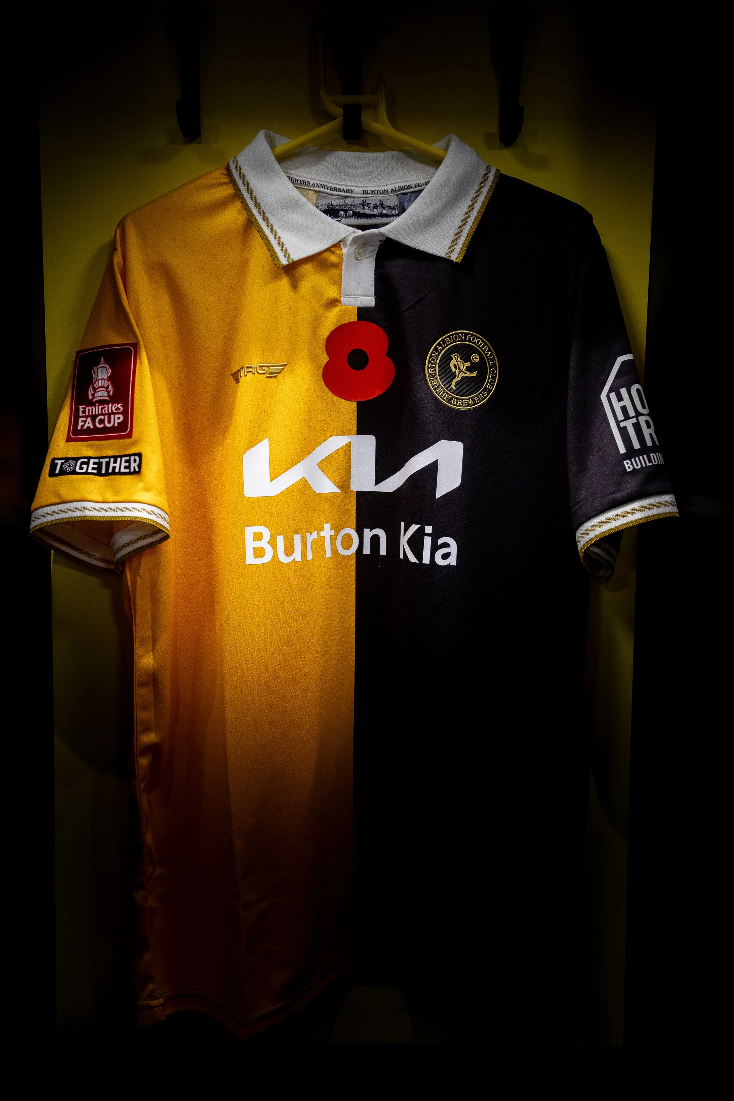 Yellow and black football jersey with Buzzard logo, sponsor logos, and a red poppy pin on the front, hanging on a yellow hanger against a yellow wall.