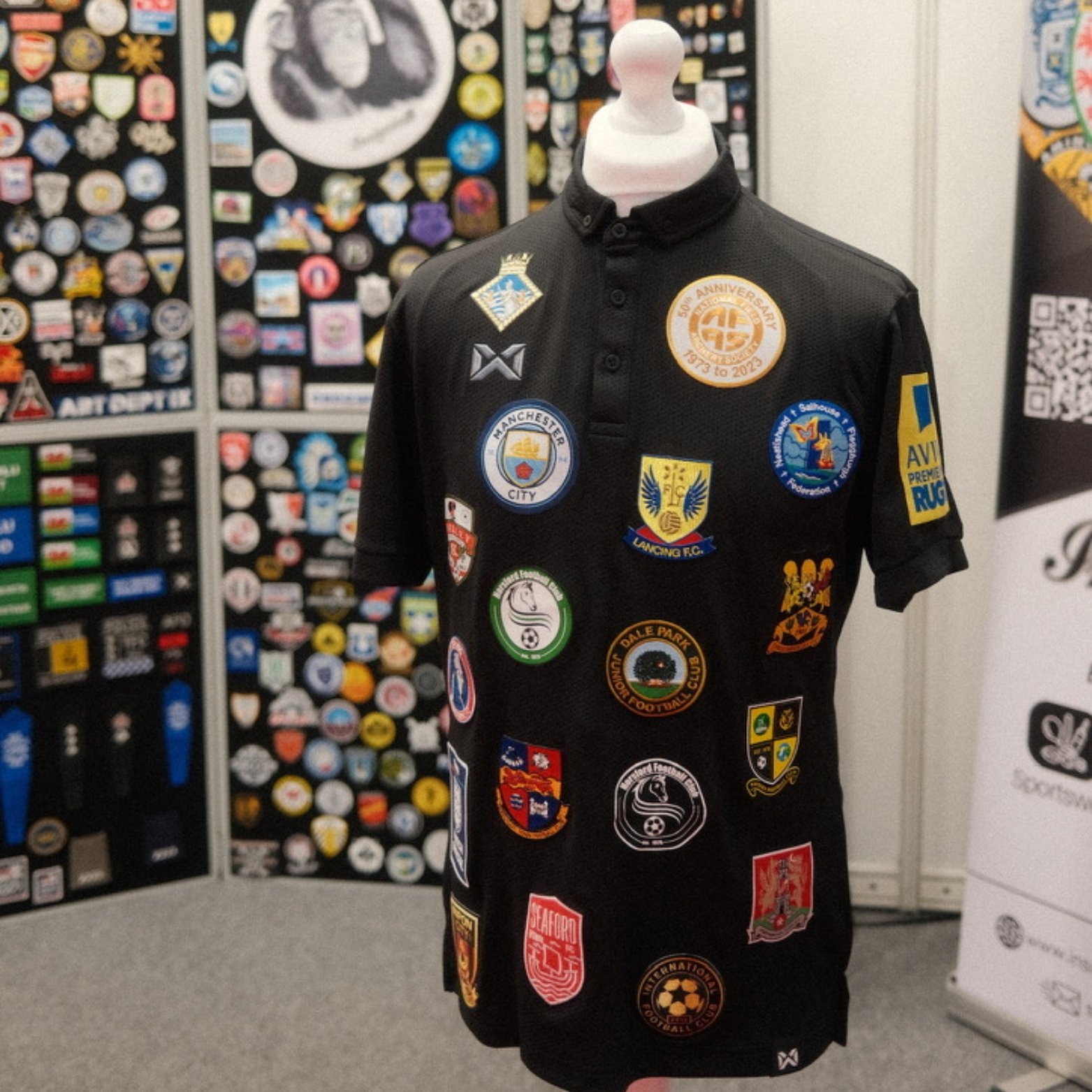 Black polo shirt covered with multiple colorful patches and badges, displayed on a mannequin with a white head. The patches include various football club logos and commemorative insignias. The background shows a display of many other patches and badges on a wall.