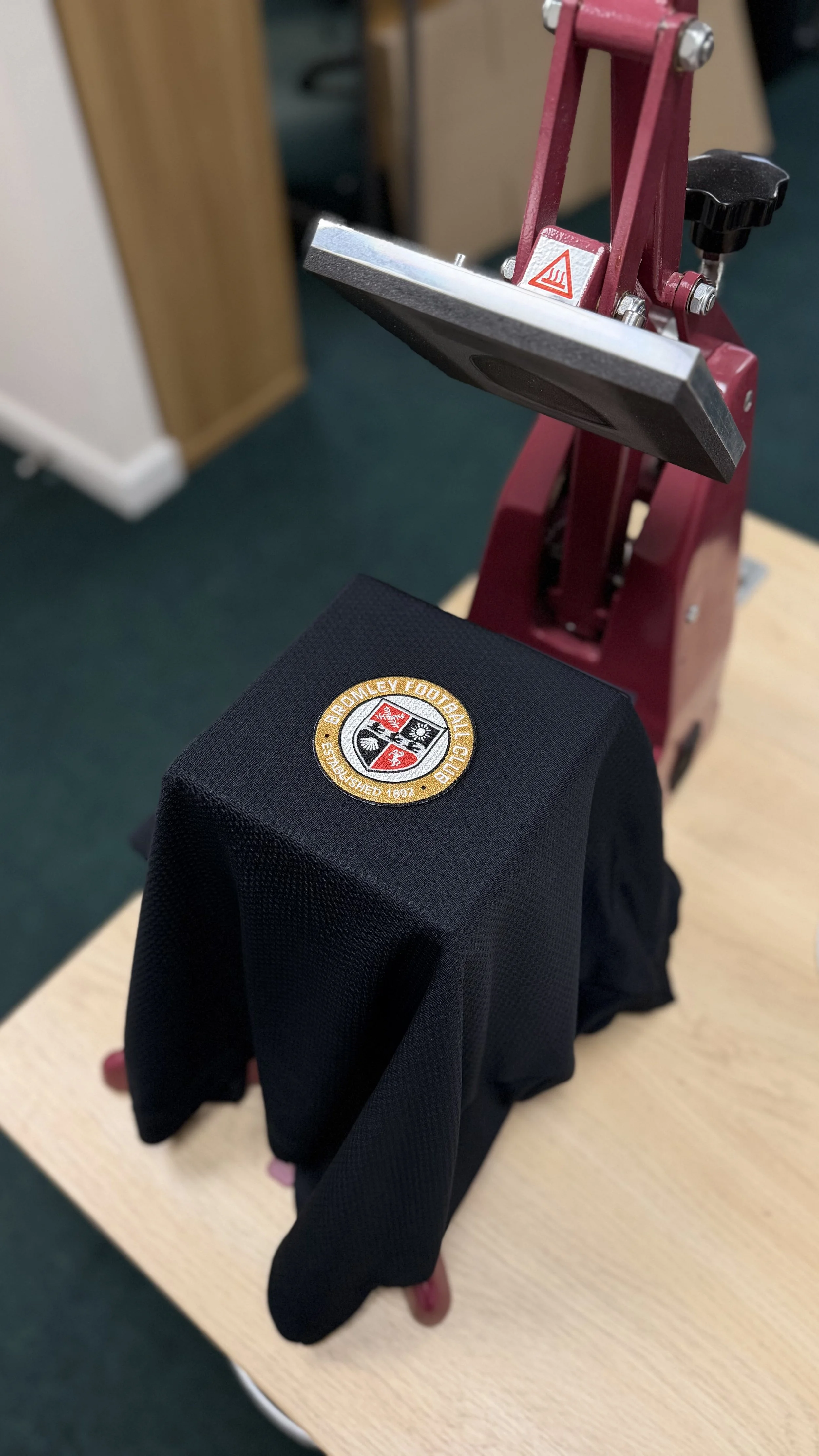 A black cloth with the Bromley Football Club crest draped over a rectangular object, with a red machine with a metallic plate attached, on a wooden surface.