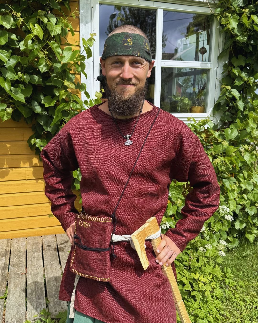 En mann kledd i middelaldersk inspirert antrekk, med en svart bandana, smilende foran en husvegg med grønne planter rundt vinduet.