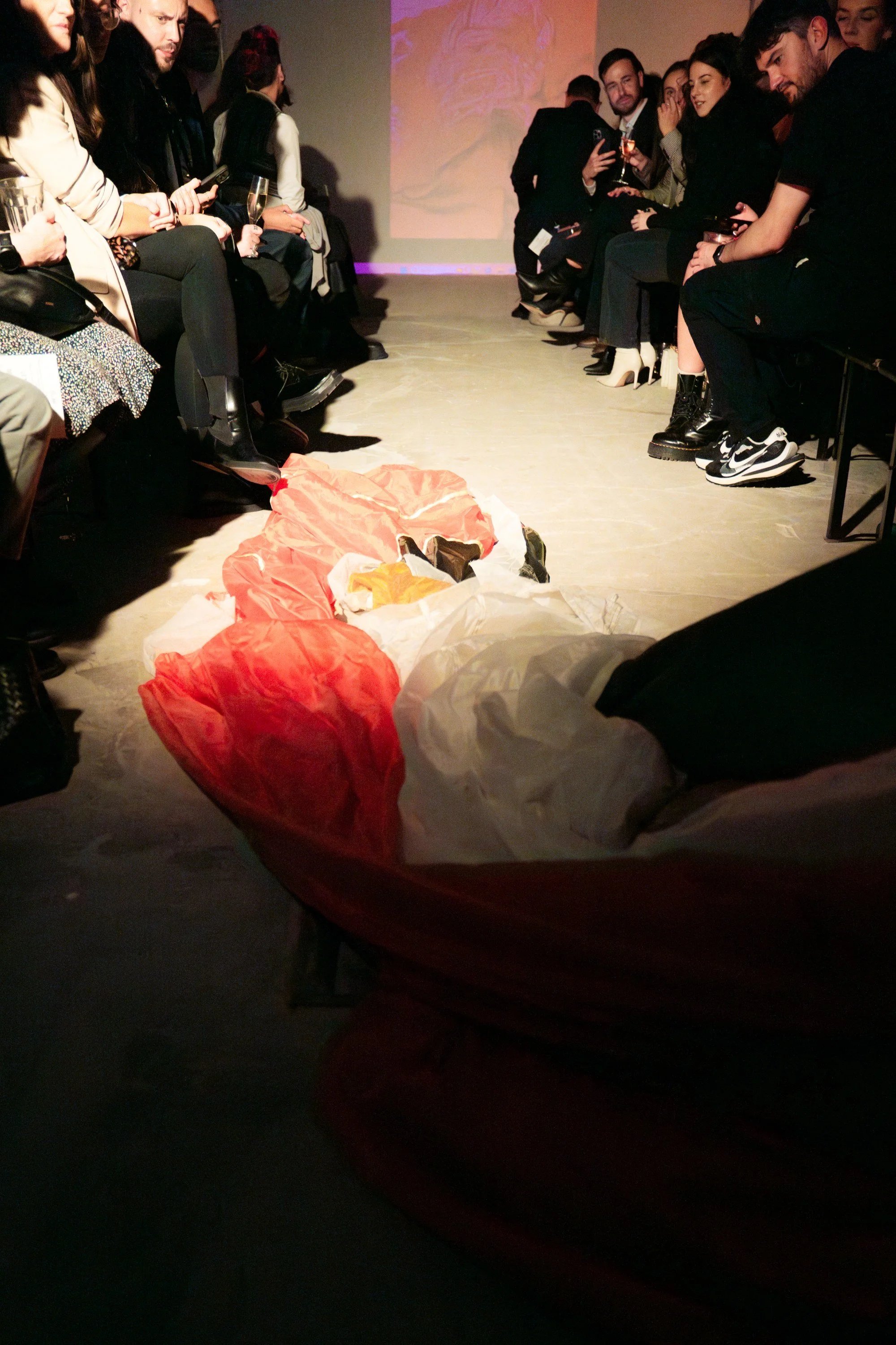 People sitting in chairs on either side of a runway, watching a person who has fallen or collapsed on the floor in the middle of the runway at an event or fashion show.